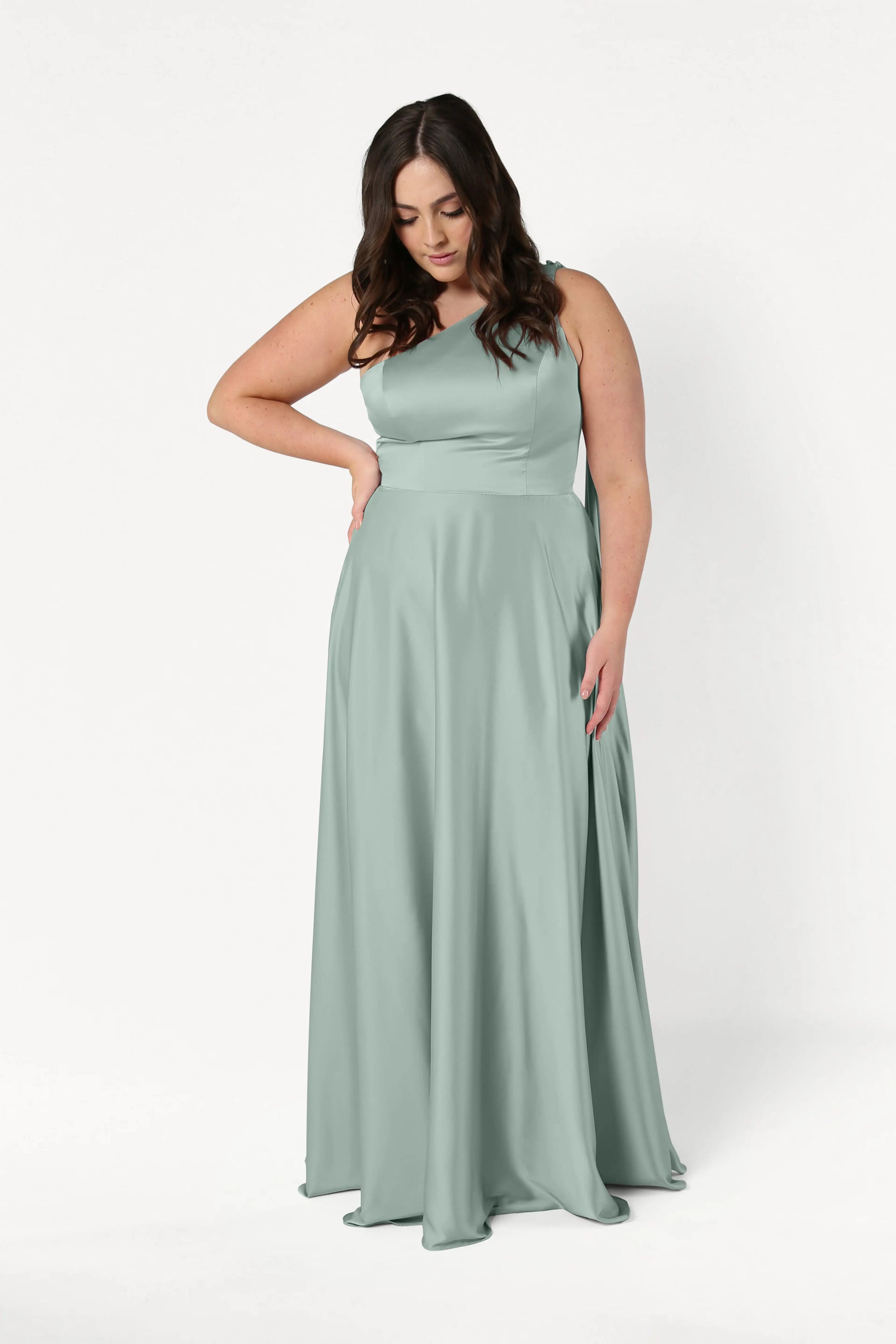 A woman with long brown hair wears the TH&TH Lyra Bridesmaid Dress in Dusty Eucalyptus Satin, standing against a white background and looking down with one hand on her hip.