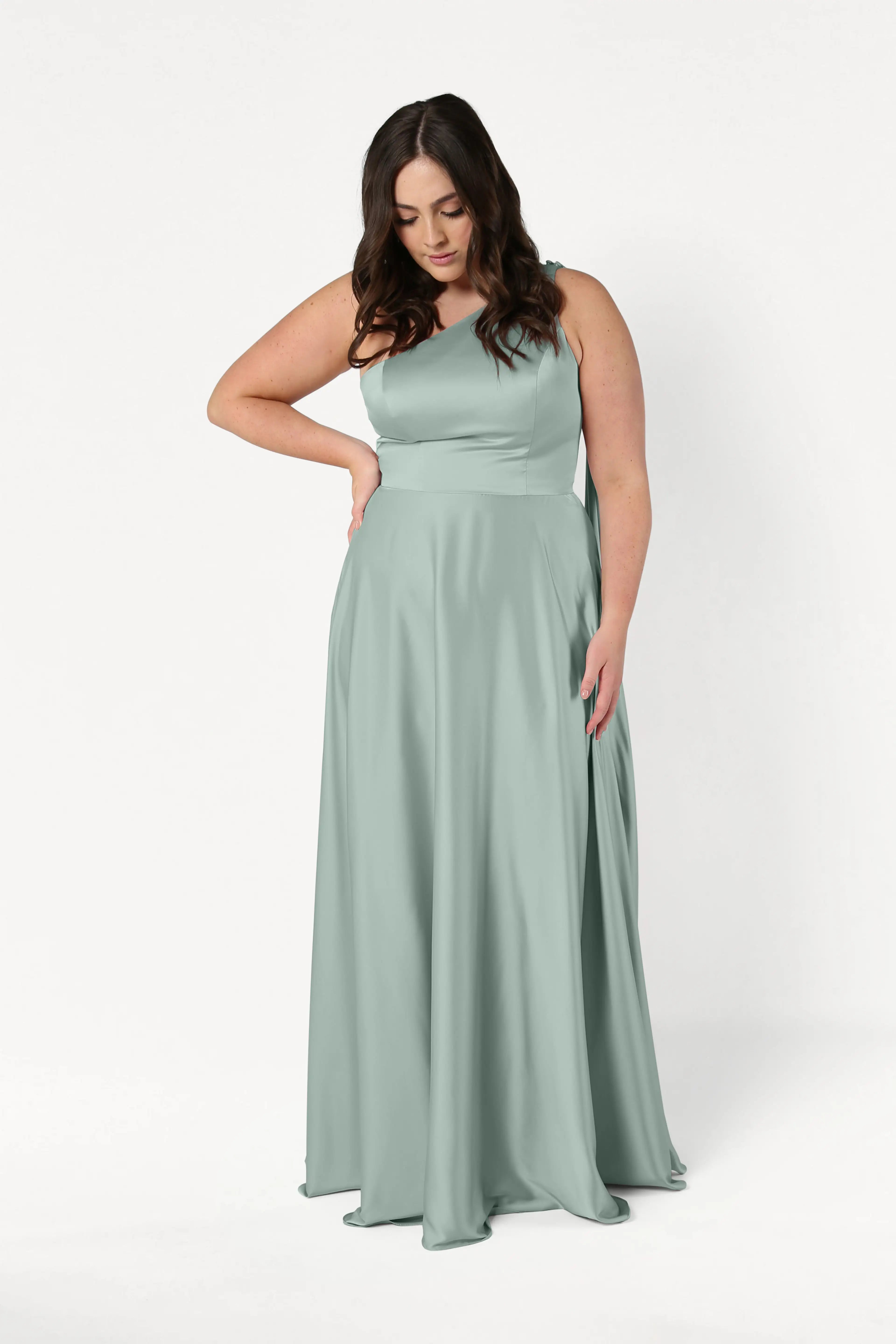 A woman with long brown hair wears the TH&TH Lyra Bridesmaid Dress in Dusty Eucalyptus Satin, standing against a white background and looking down with one hand on her hip.