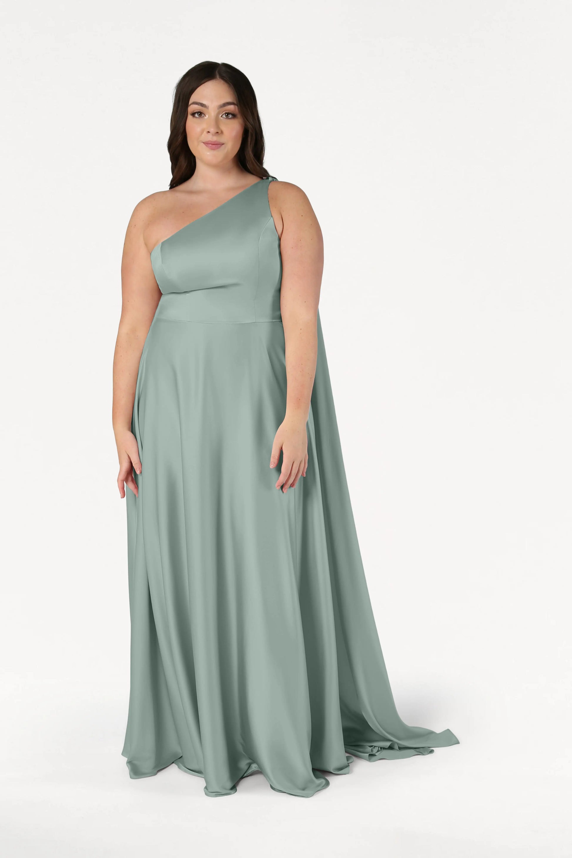 A woman models the TH&TH Lyra Bridesmaid Dress in Dusty Eucalyptus Satin—a size-inclusive, floor-length, one-shoulder gown—against a plain white background, looking at the camera with a neutral expression.