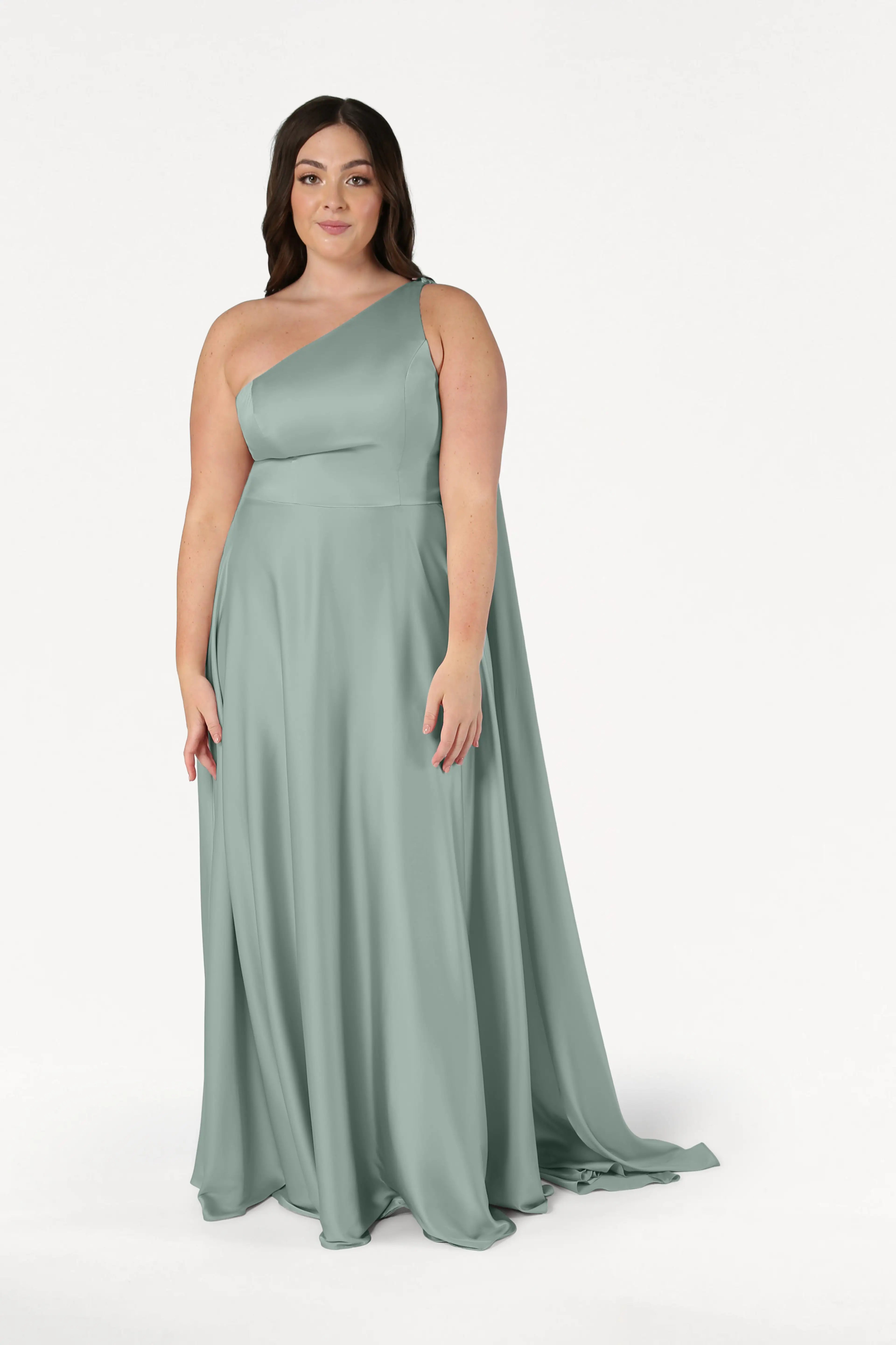 A woman models the TH&TH Lyra Bridesmaid Dress in Dusty Eucalyptus Satin—a size-inclusive, floor-length, one-shoulder gown—against a plain white background, looking at the camera with a neutral expression.