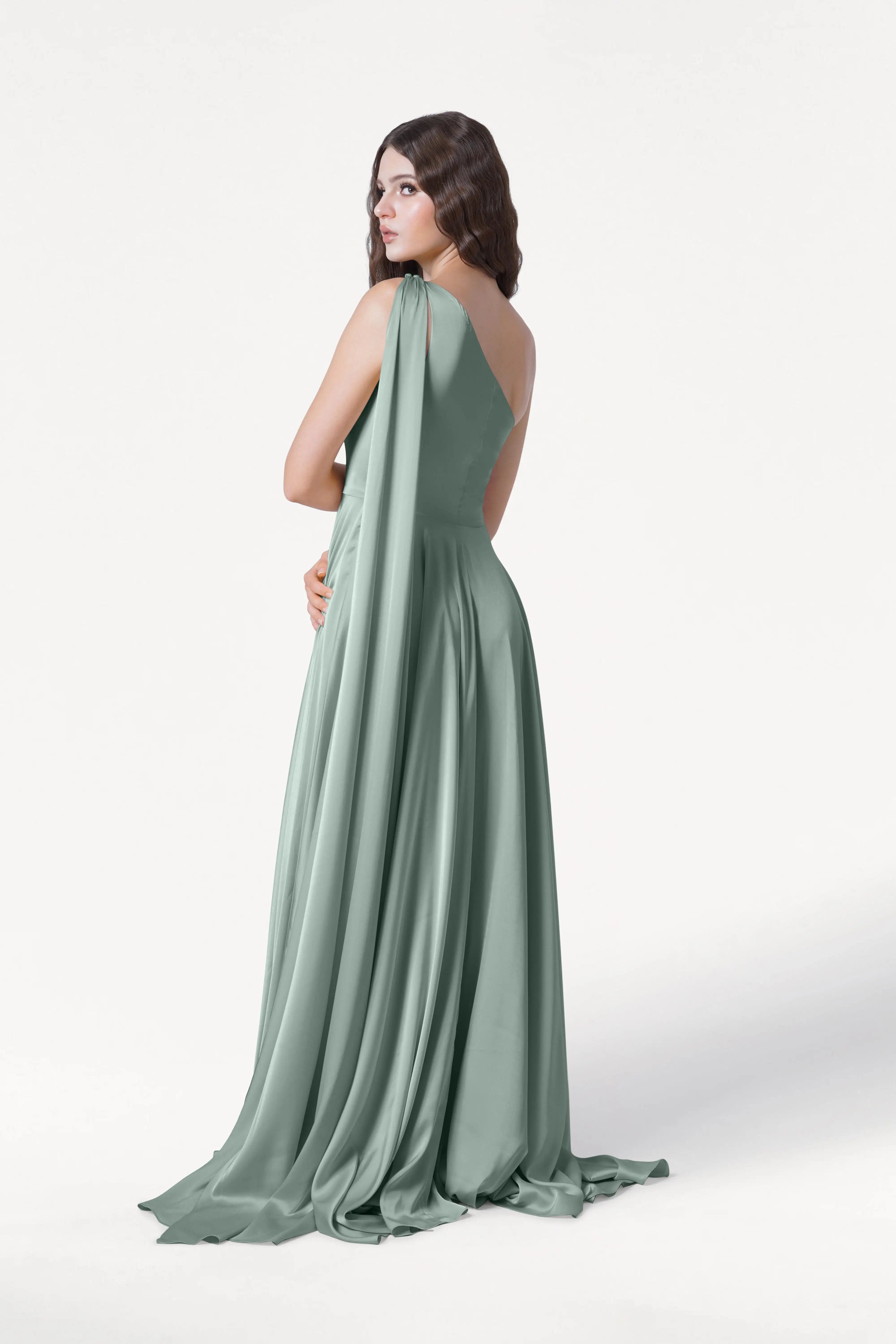 A woman with long brown hair models the Lyra Bridesmaid Dress in Dusty Eucalyptus Satin by TH&TH, a size-inclusive, floor-length gown with draped shoulders and an elegant train, standing slightly turned against a white background.