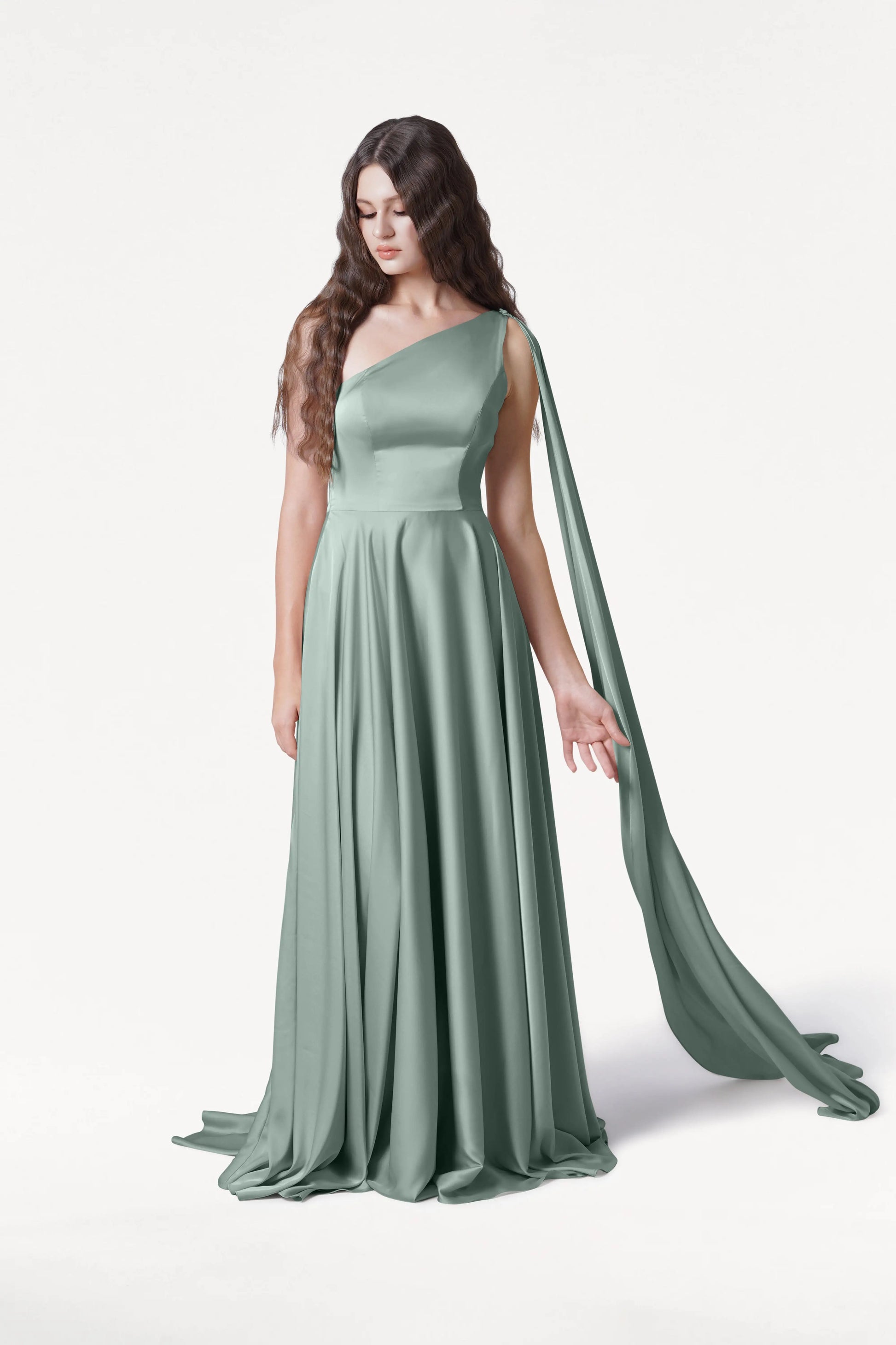 A woman with long wavy brown hair wears the Lyra Bridesmaid Dress in Dusty Eucalyptus Satin by TH&TH, a flowing one-shoulder gown with a draped sash. She stands against a white background, looking down with one hand gently extended.