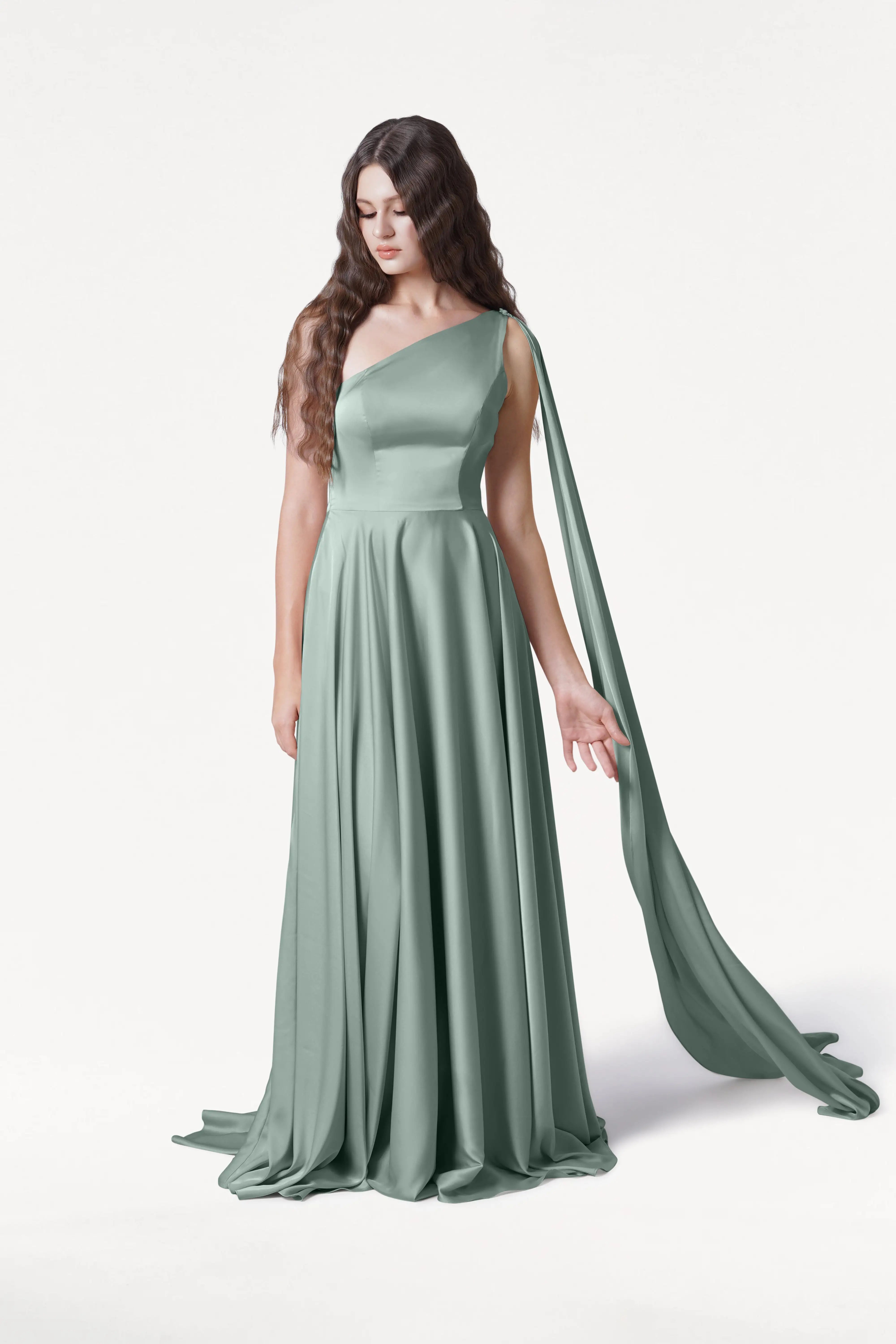 A woman with long wavy brown hair wears the Lyra Bridesmaid Dress in Dusty Eucalyptus Satin by TH&TH, a flowing one-shoulder gown with a draped sash. She stands against a white background, looking down with one hand gently extended.