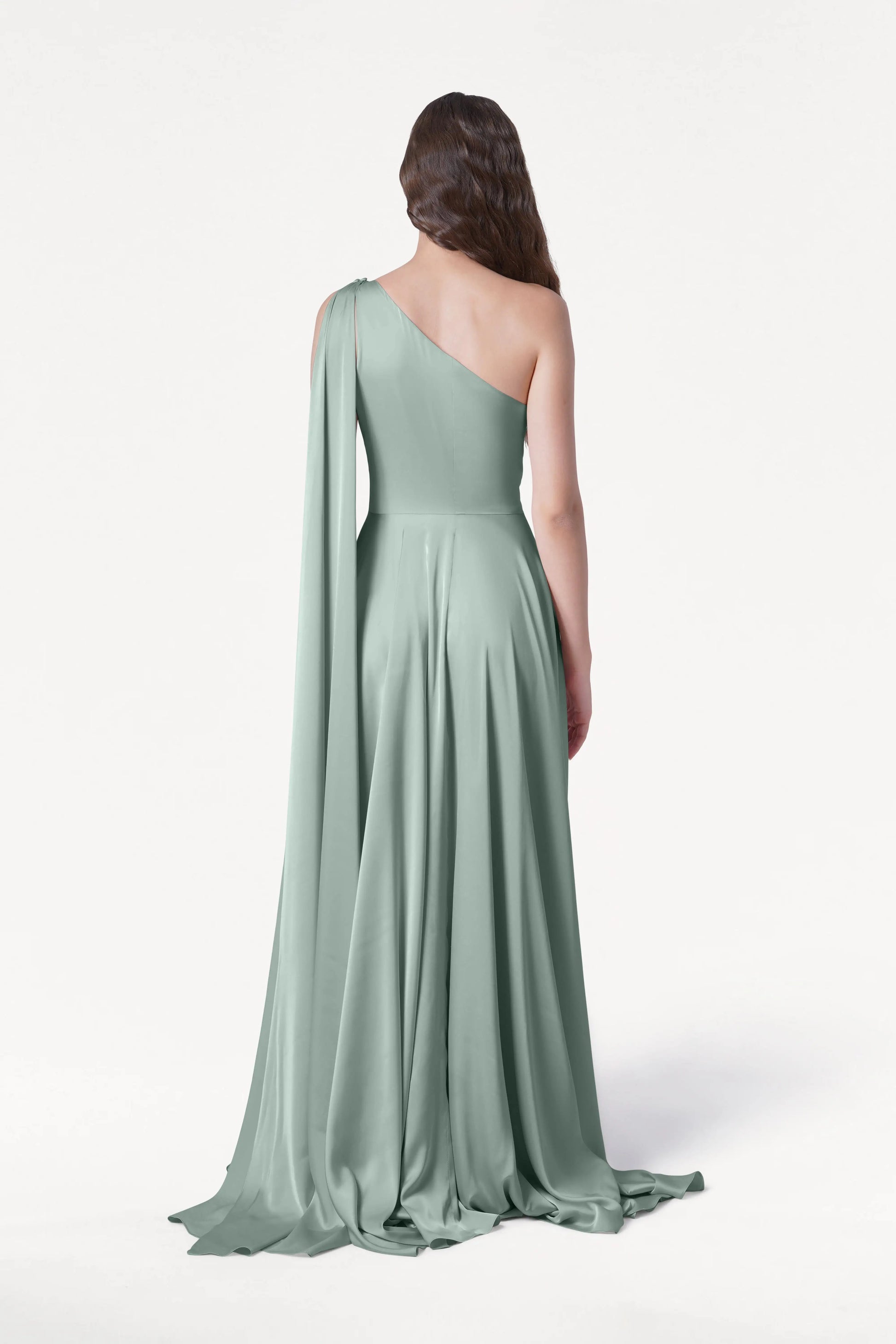 A woman in the TH&TH Lyra Bridesmaid Dress in Dusty Eucalyptus Satin, featuring a long, flowing silhouette and an asymmetrical one-shoulder design, stands facing away against a plain white background.