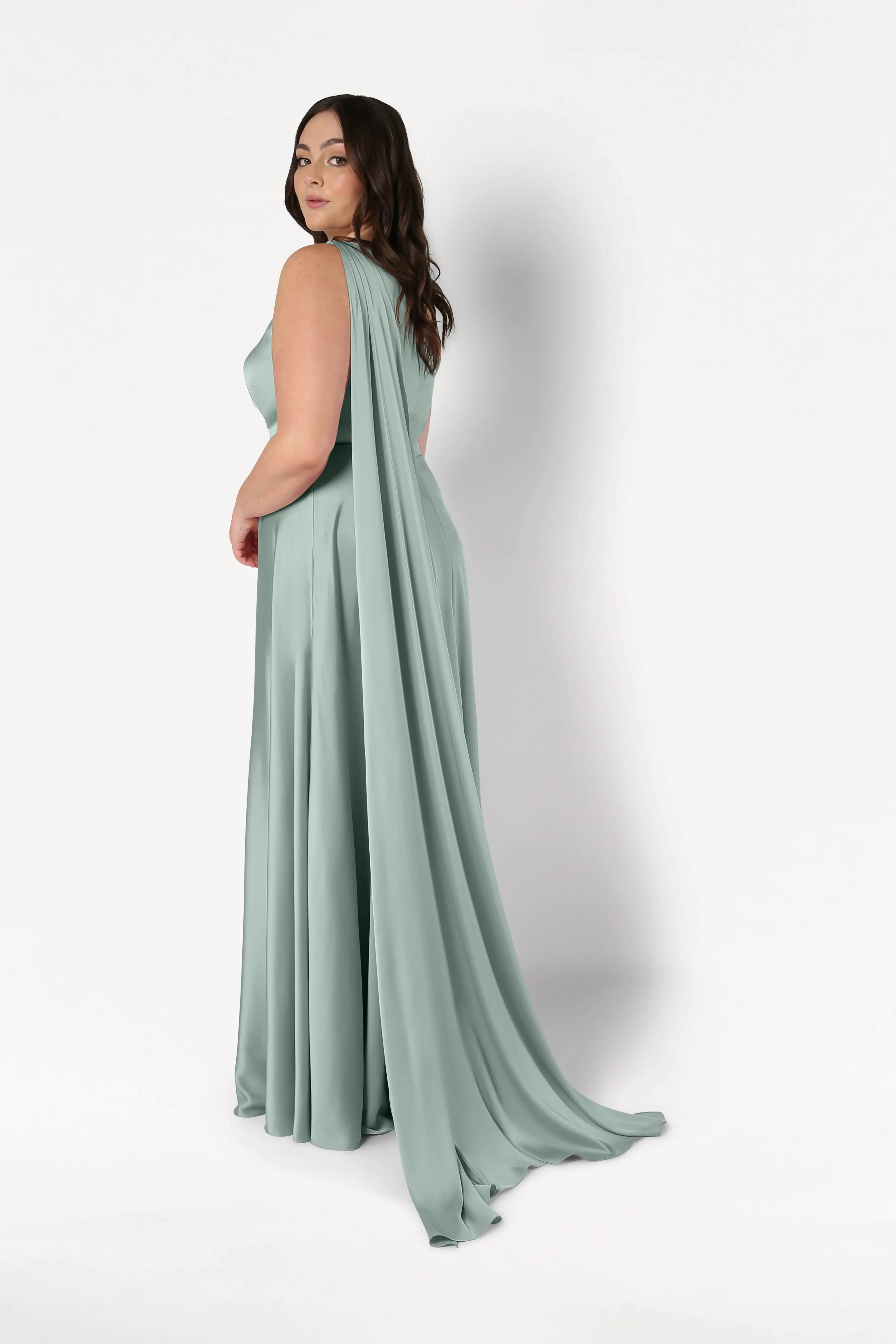 A woman in the TH&TH Lyra Bridesmaid Dress in Dusty Eucalyptus Satin, a size-inclusive, floor-length gown with a flowing cape, stands turned slightly to the side against a plain white background, looking over her shoulder.