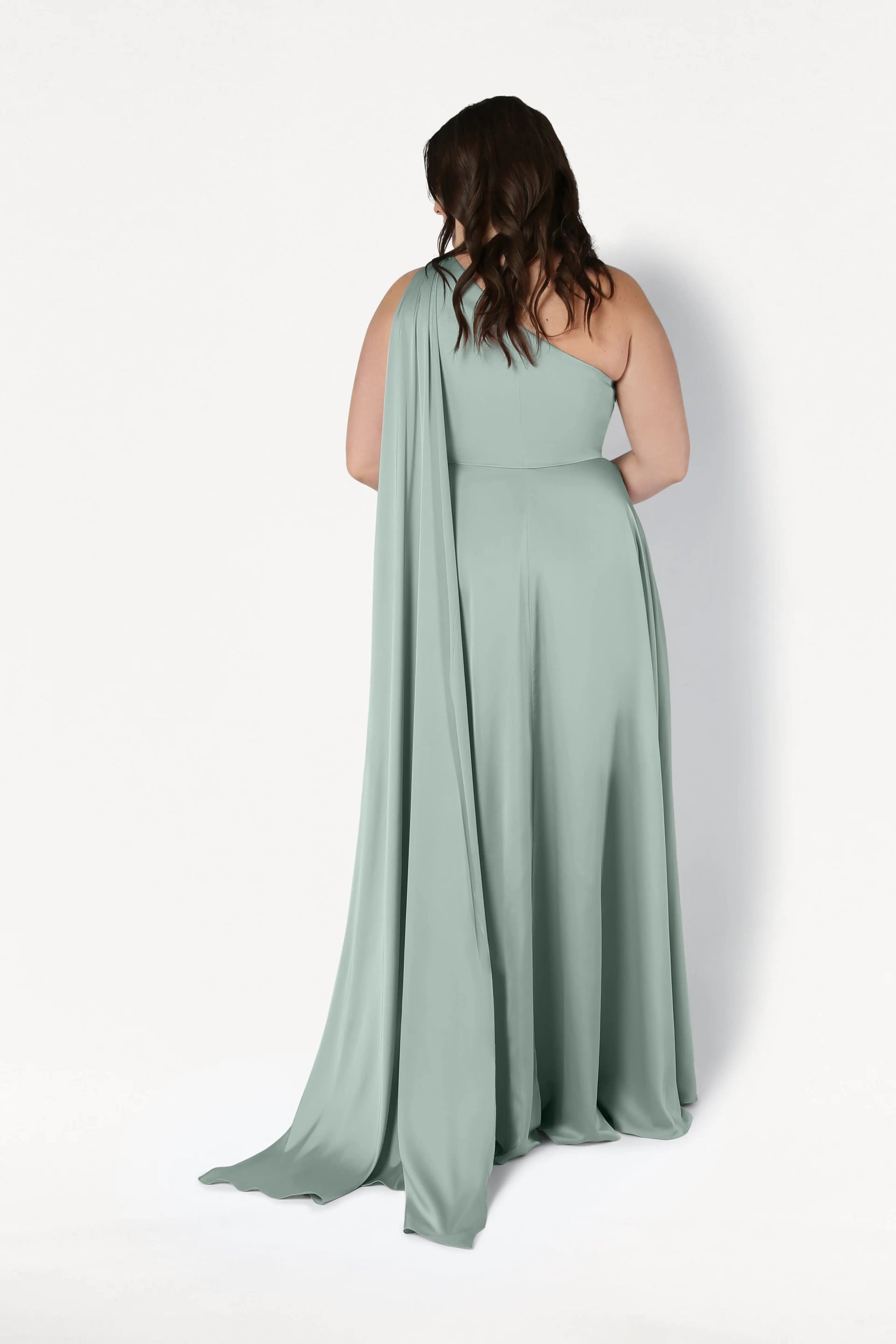 A woman with wavy brown hair faces away in the TH&TH Lyra Bridesmaid Dress in Dusty Eucalyptus Satin, a size-inclusive, floor-length, one-shoulder gown with a flowing drape, set against a plain white background.