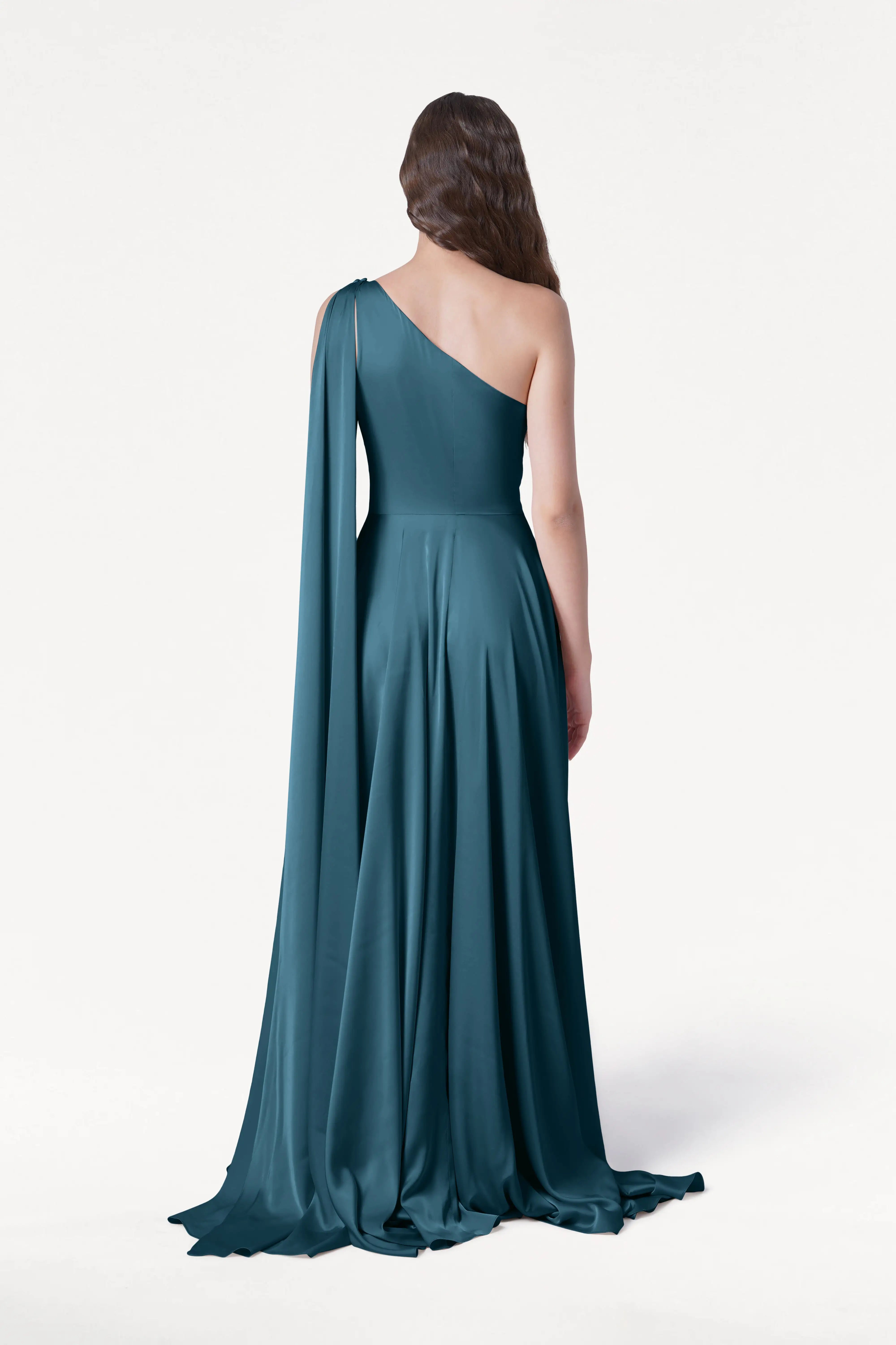 A person with long wavy hair stands facing away against a plain white background, wearing the TH&TH Lyra Bridesmaid Dress in Emerald Dawn Satin—a modern, floor-length one-shoulder gown with a draped sash.