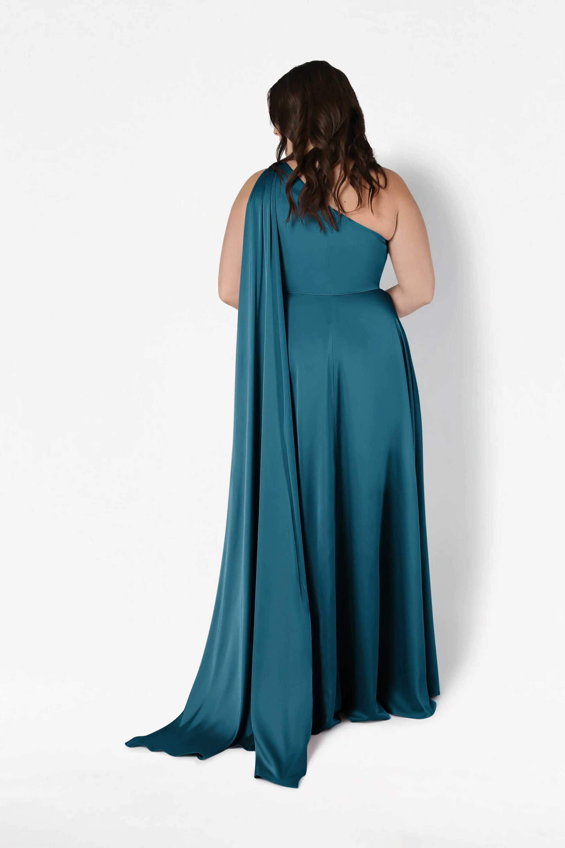 A woman with wavy brown hair stands facing away, wearing the TH&TH Lyra Bridesmaid Dress in Emerald Dawn Satin—a teal, one-shoulder gown with a long, flowing drape—set against a white background.