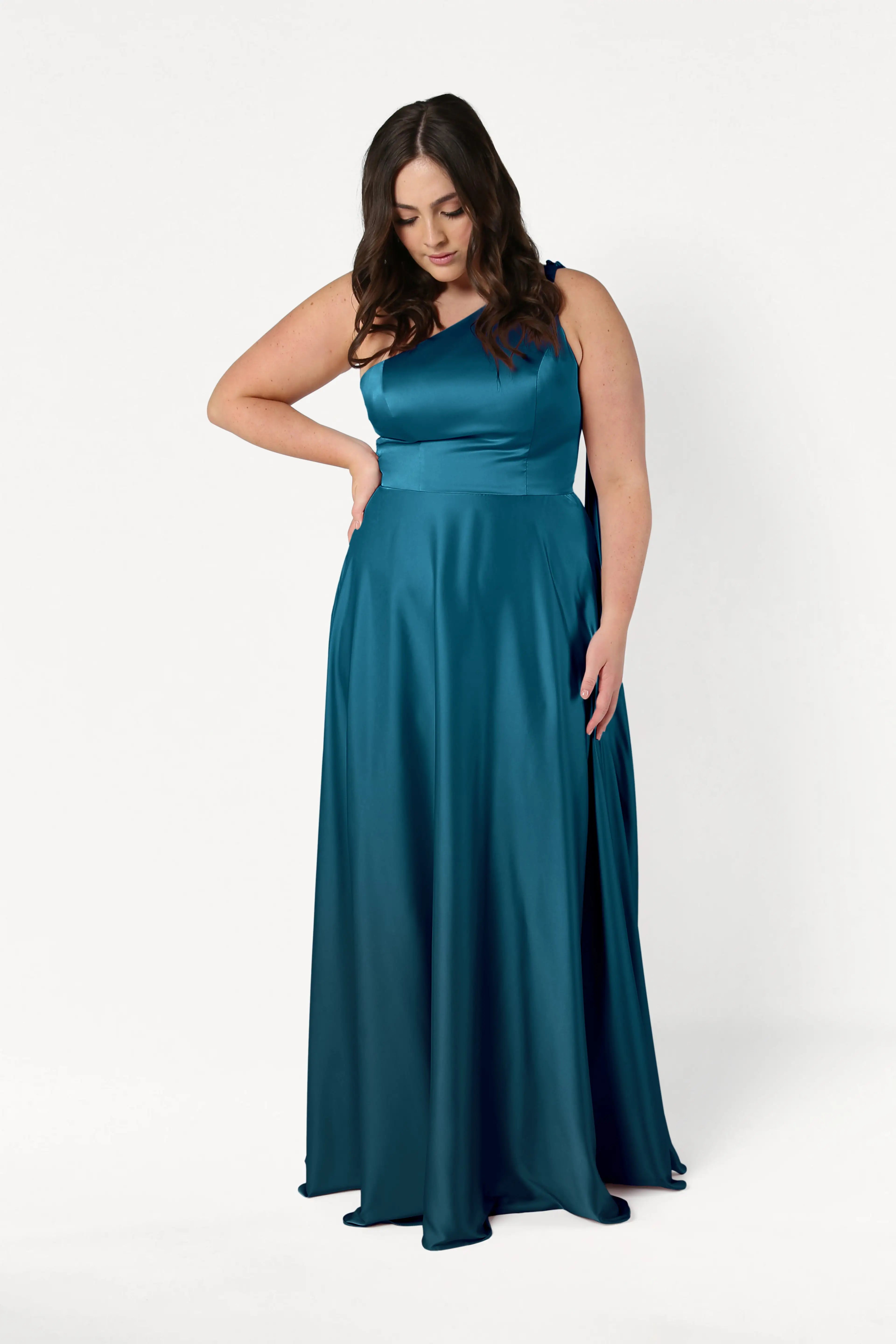 A woman models the TH&TH Lyra Bridesmaid Dress in Emerald Dawn Satin, a size-inclusive, one-shoulder gown, standing against a plain white background with one hand on her hip and looking down.