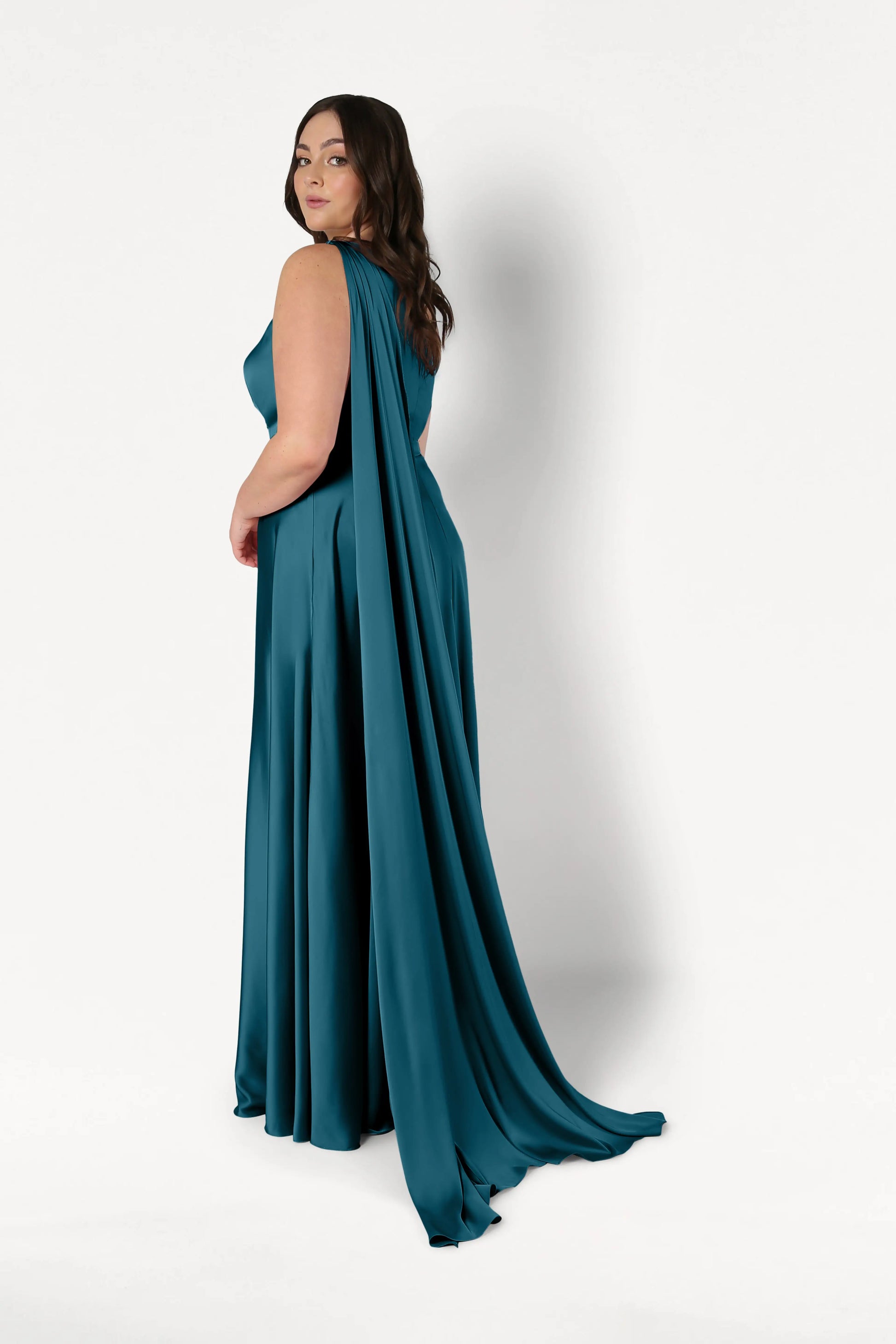 A woman with long brown hair models the TH&TH Lyra Bridesmaid Dress in Emerald Dawn Satin, featuring a dramatic cape detail, as she stands against a plain white background and looks over her shoulder.
