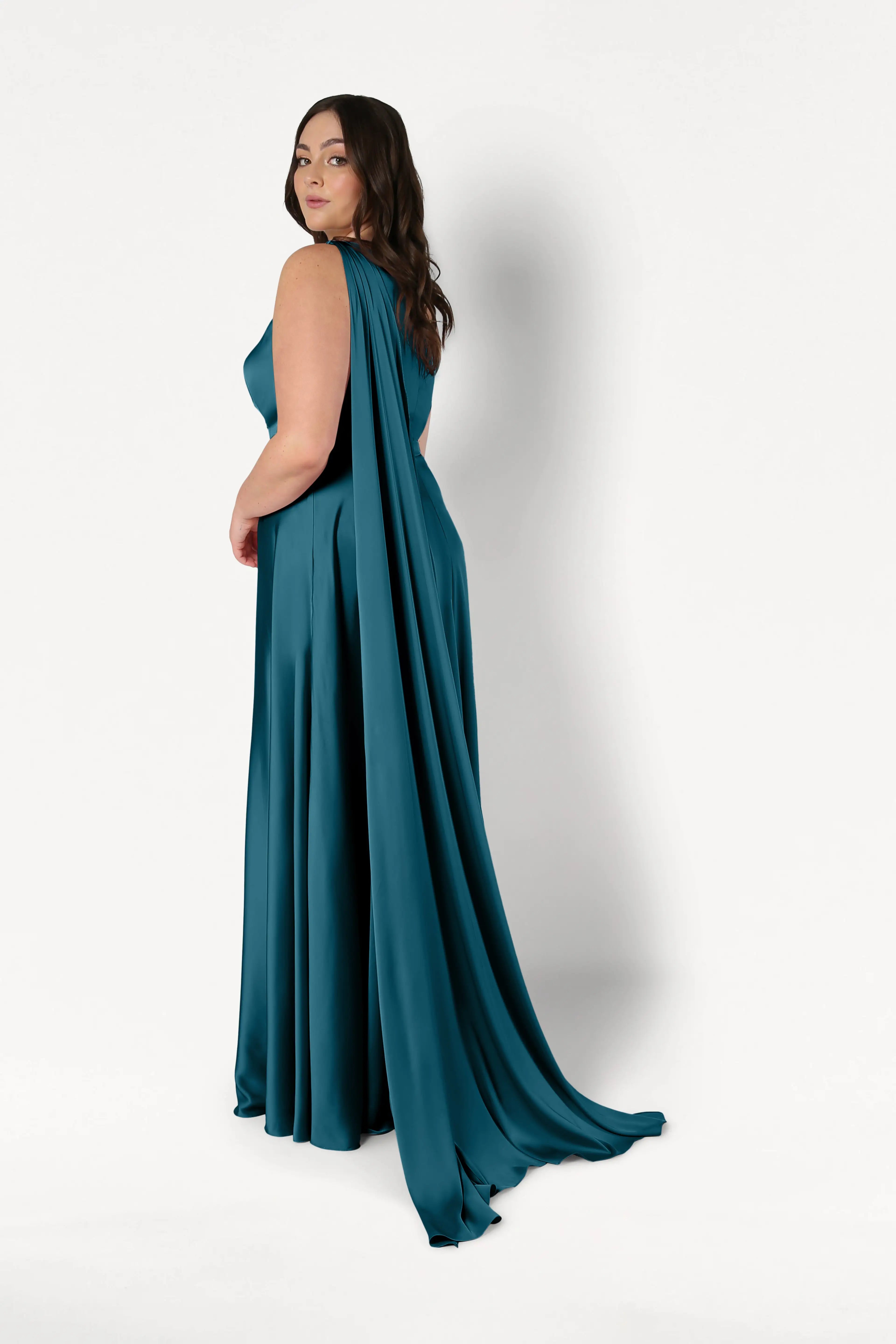 A woman with long brown hair models the TH&TH Lyra Bridesmaid Dress in Emerald Dawn Satin, featuring a dramatic cape detail, as she stands against a plain white background and looks over her shoulder.