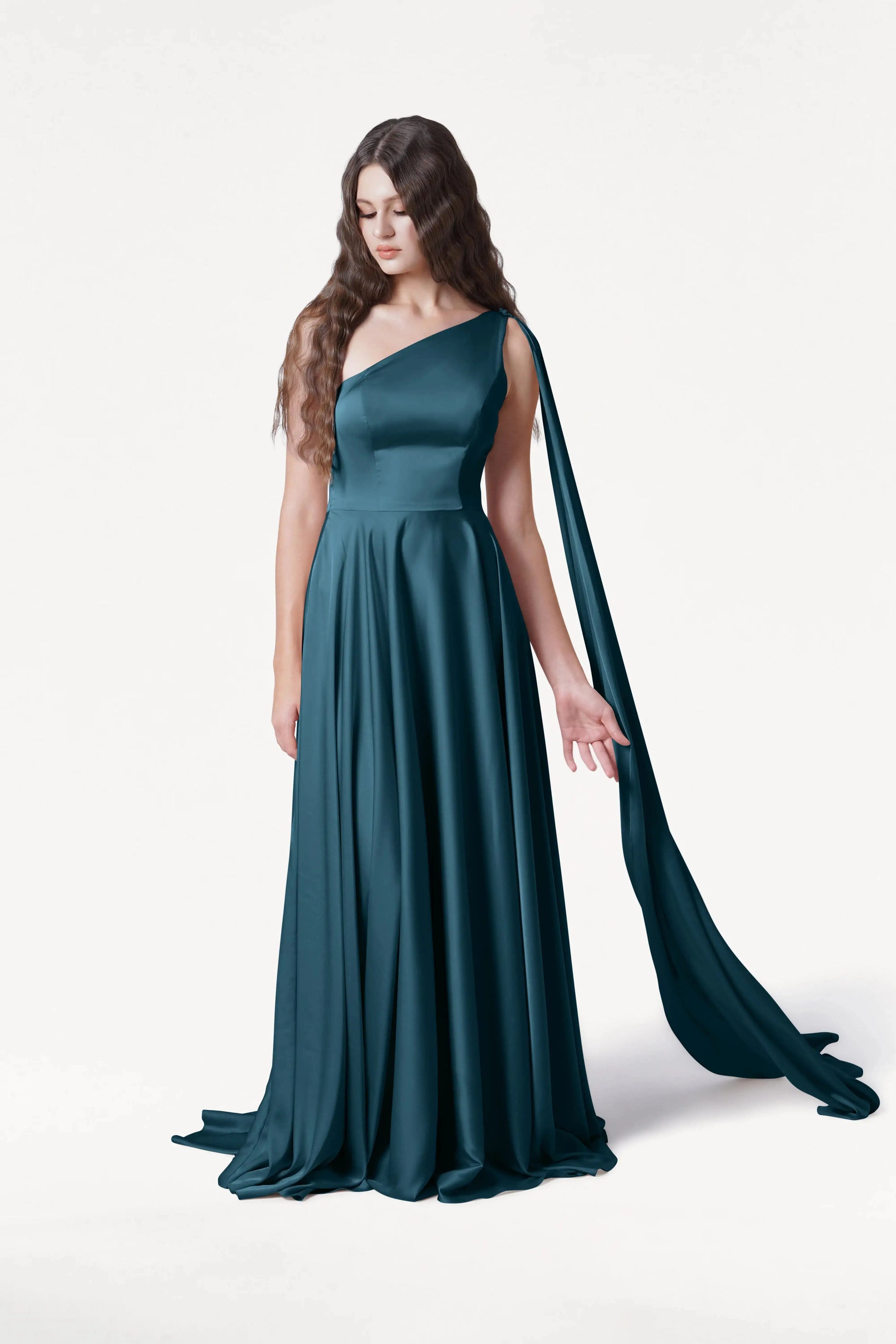 A woman with long wavy brown hair models the Lyra Bridesmaid Dress in Emerald Dawn Satin by TH&TH—a size-inclusive, floor-length, one-shoulder teal gown with a flowing scarf detail—against a plain white background.