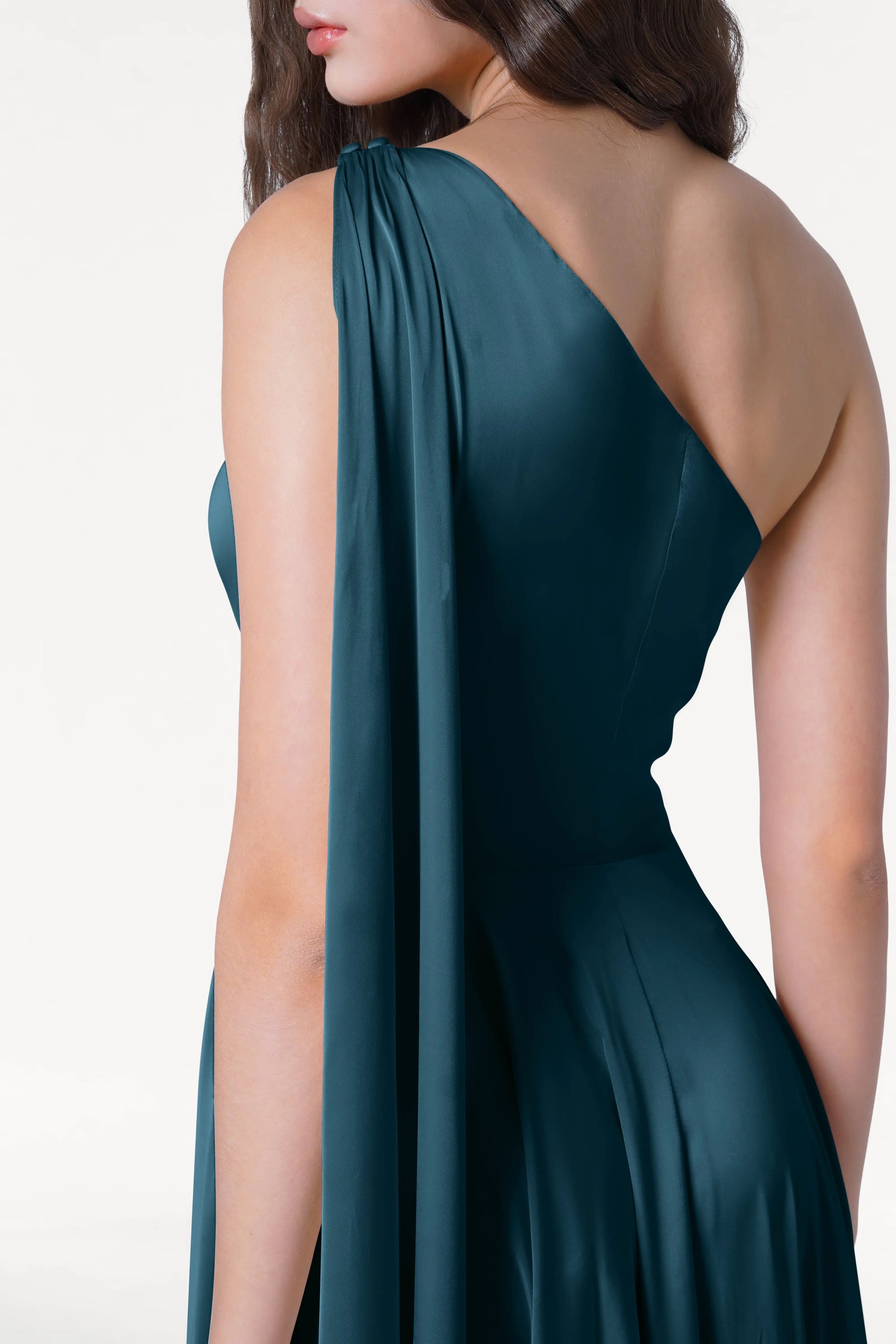 A woman models the Lyra Bridesmaid Dress in Emerald Dawn Satin by TH&TH, a sleeveless, one-shoulder gown with draped satin detail. She stands against a white background with side-parted hair, showcasing an elegant, modern bridal look.
