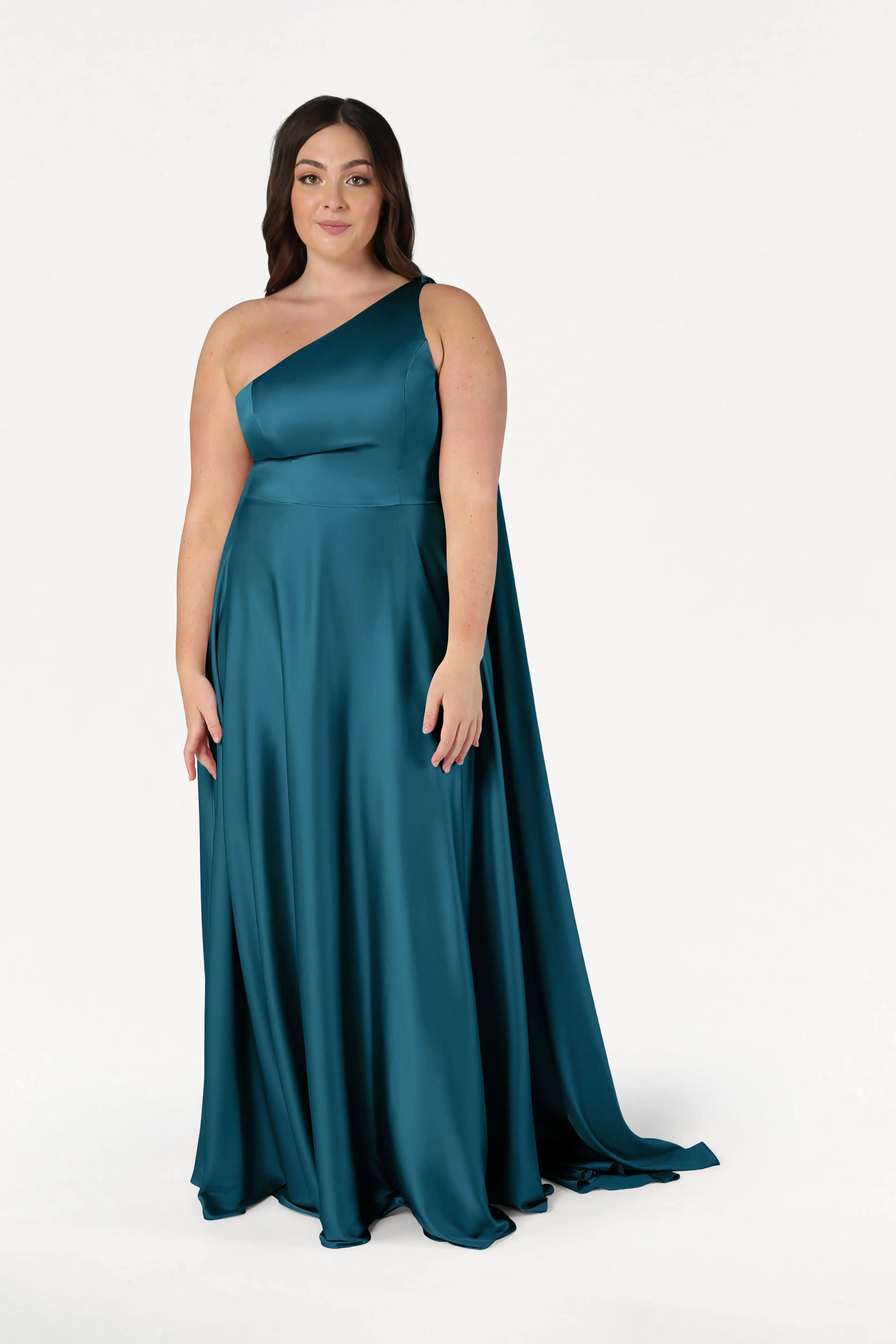 A woman stands against a white background wearing the TH&TH Lyra Bridesmaid Dress in Emerald Dawn Satin—an elegant, modern, floor-length, one-shoulder gown—looking forward with a gentle expression.
