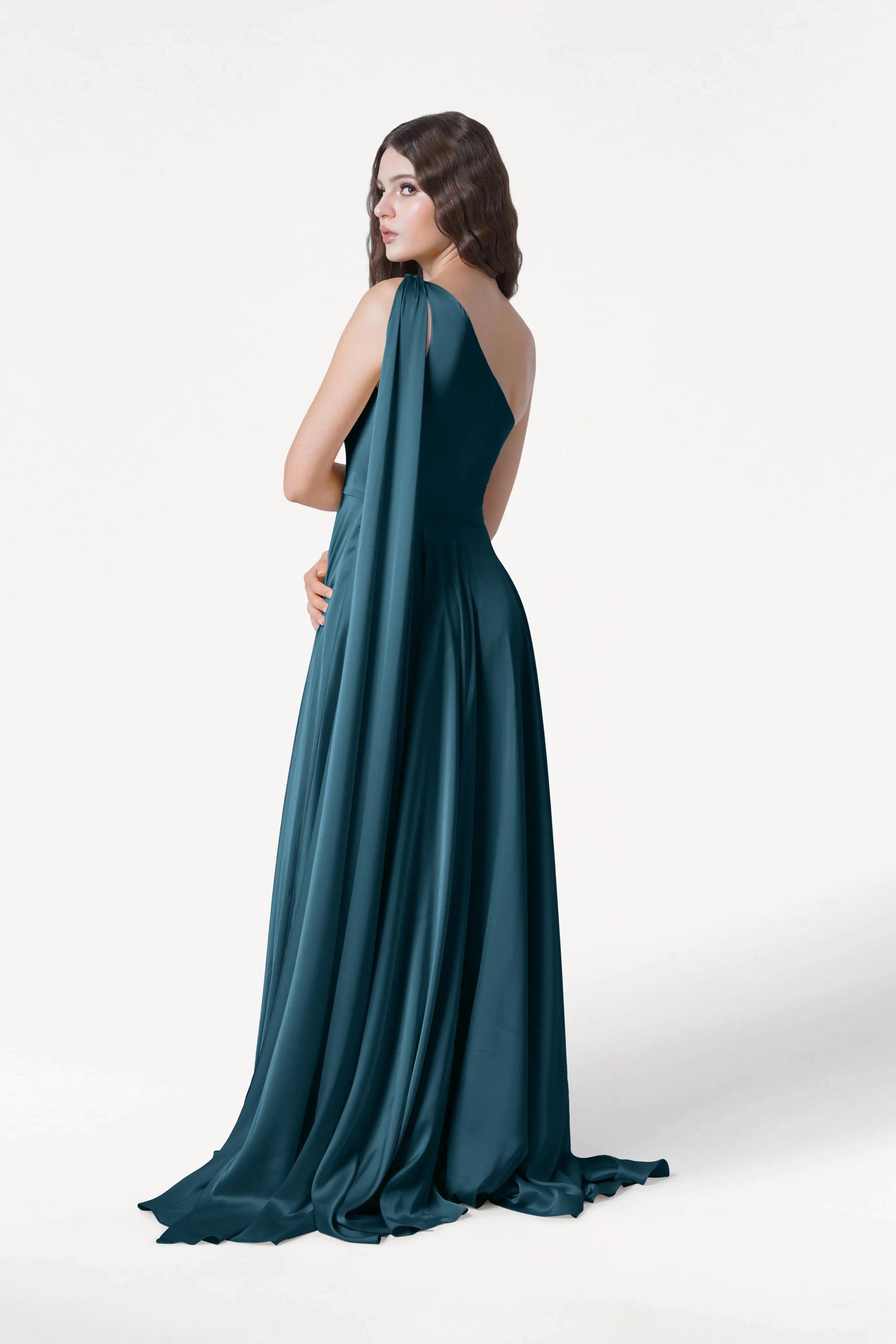 A woman with long wavy hair models the TH&TH Lyra Bridesmaid Dress in Emerald Dawn Satin, a floor-length, size-inclusive gown featuring draped fabric over one shoulder, set against a plain white background.