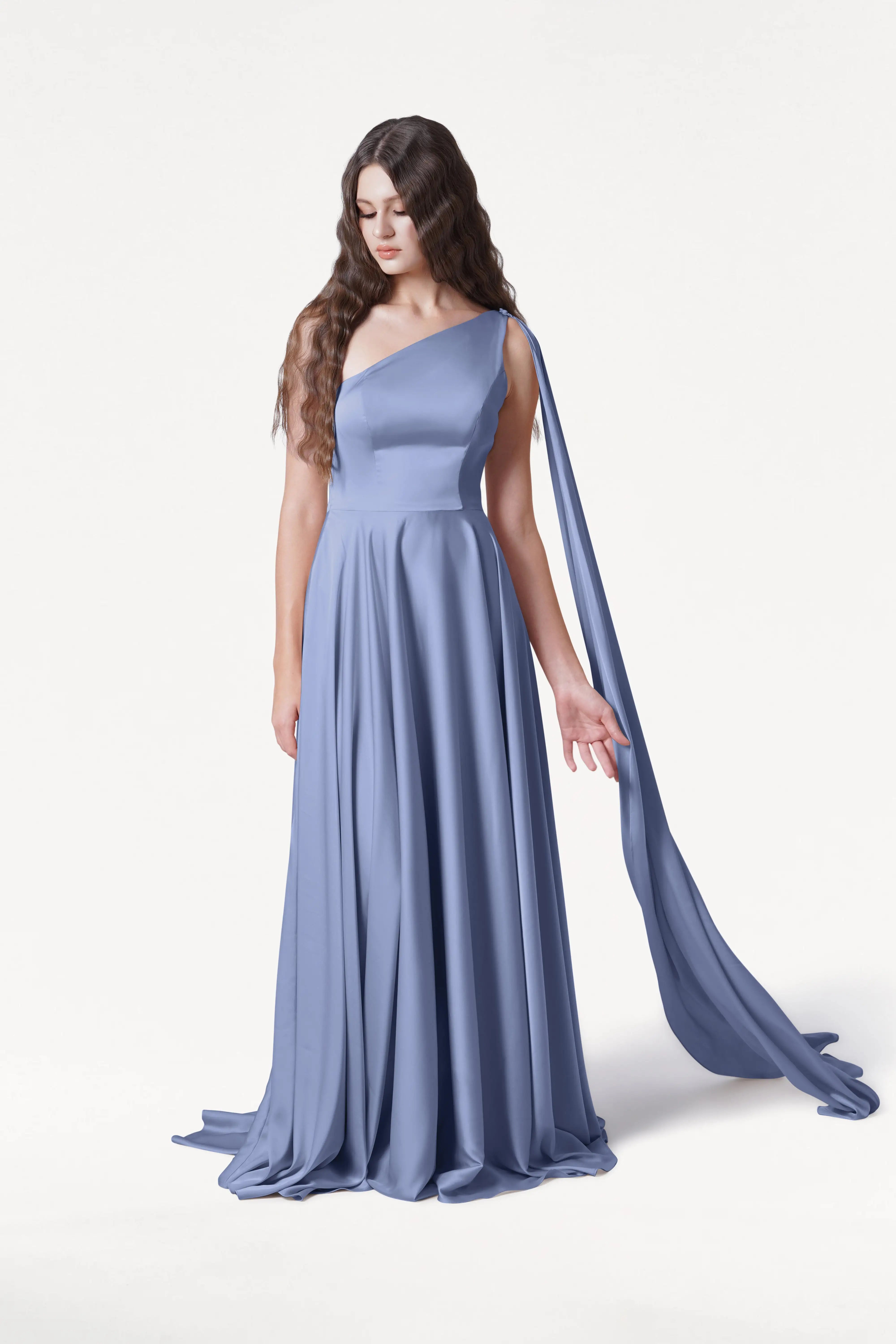 A woman with long, wavy brown hair wears the size-inclusive Lyra Bridesmaid Dress in French Blue Satin by TH&TH, featuring a one-shoulder design and draped sash. She stands gracefully against a plain white background, looking down with one arm extended.