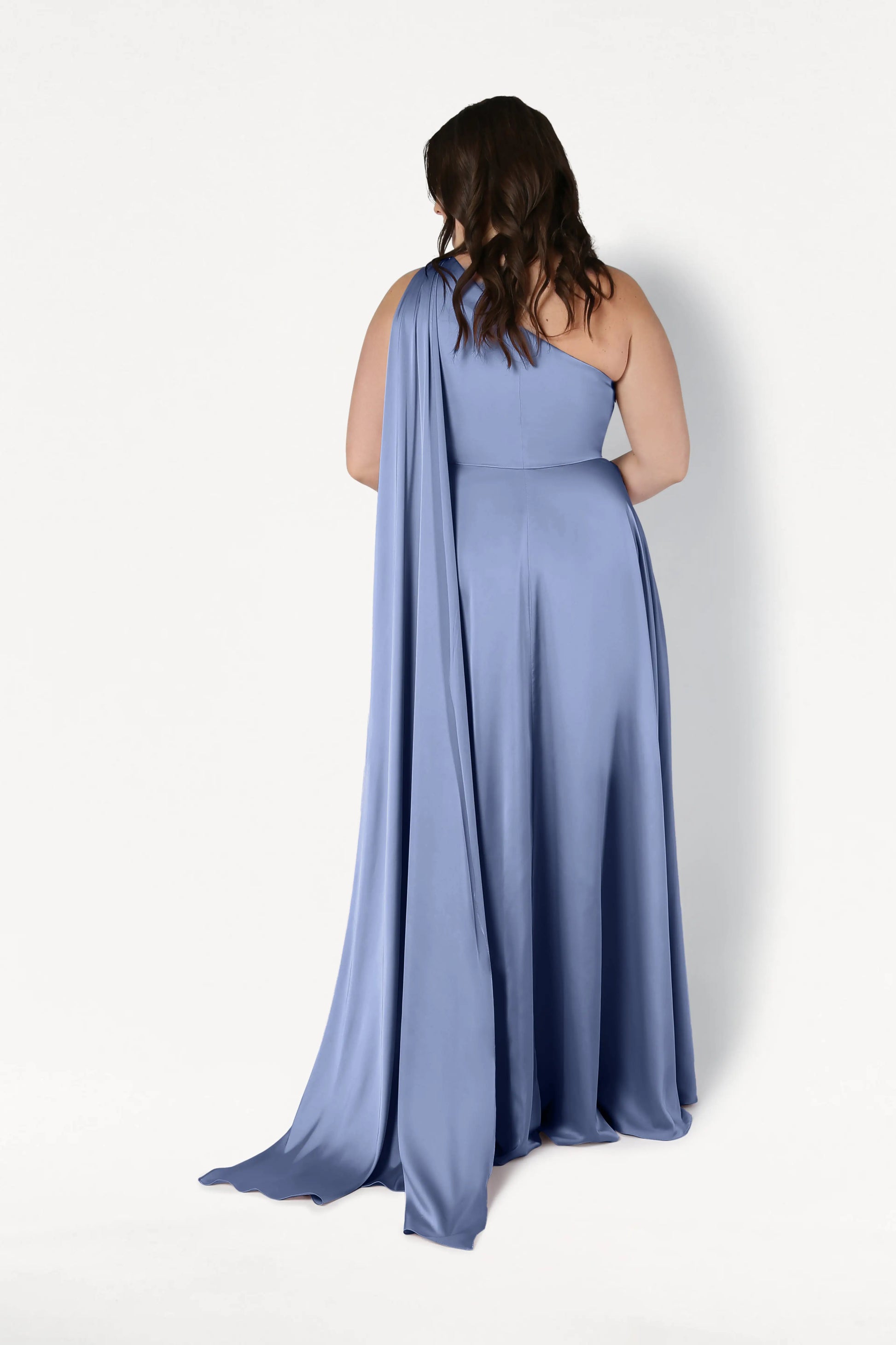A woman with wavy brown hair wears the TH&TH Lyra Bridesmaid Dress in French Blue Satin, a floor-length, one-shoulder gown with a long drape, standing with her back to the camera against a plain white background.