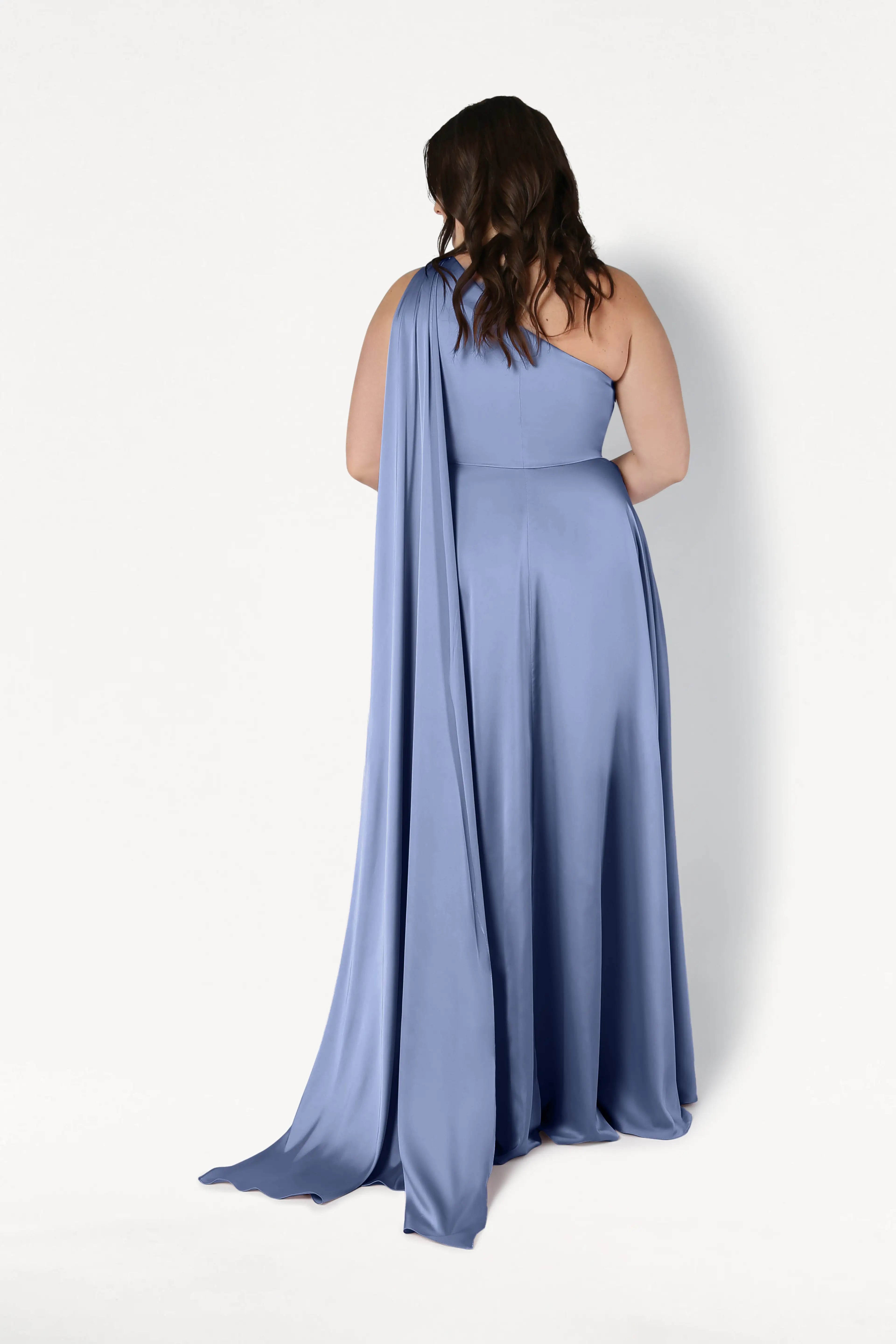 A woman with wavy brown hair wears the TH&TH Lyra Bridesmaid Dress in French Blue Satin, a floor-length, one-shoulder gown with a long drape, standing with her back to the camera against a plain white background.