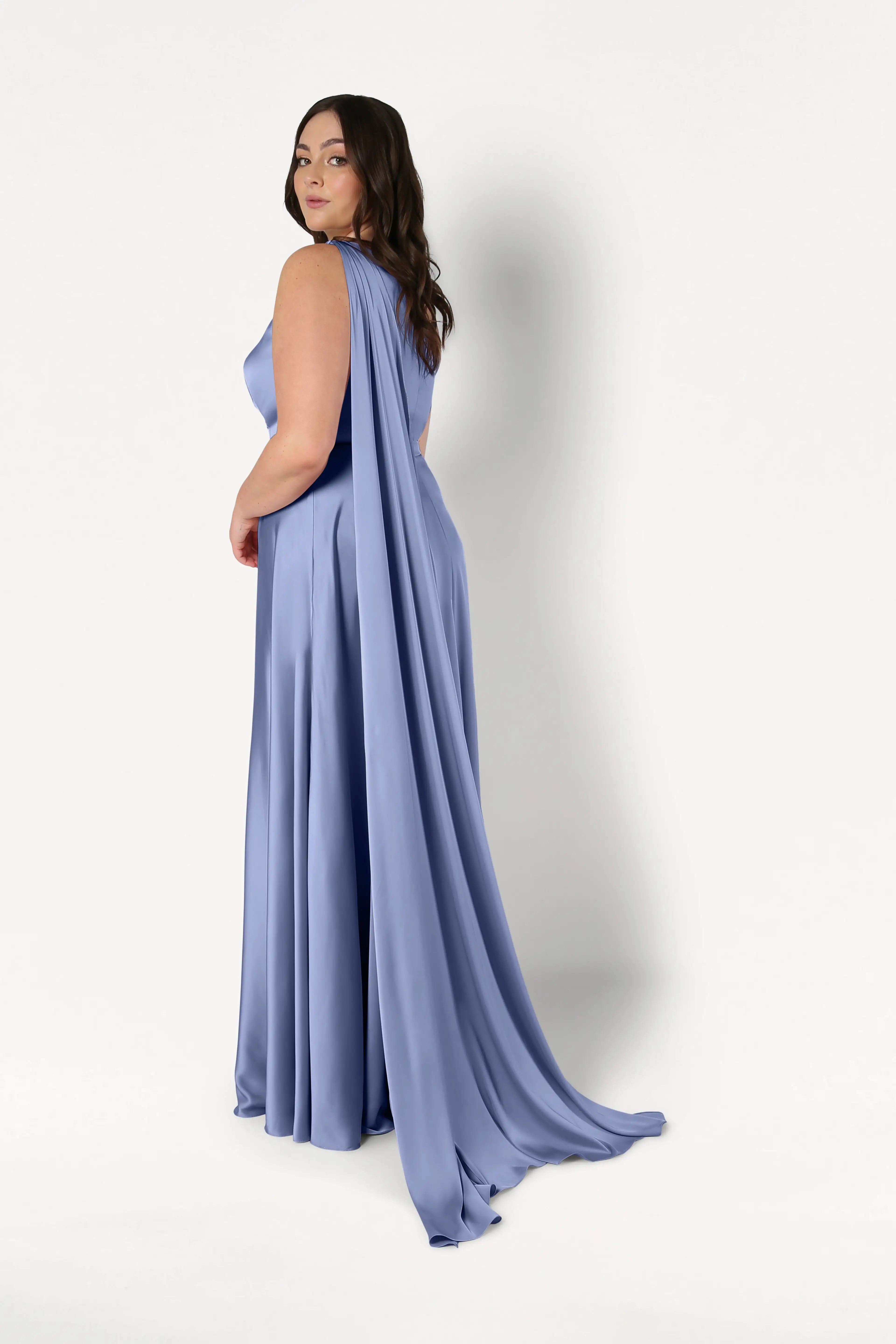A woman poses in the TH&TH Lyra Bridesmaid Dress in French Blue Satin, featuring a floor-length silhouette and a long draped cape, as she looks over her shoulder against a plain white background.