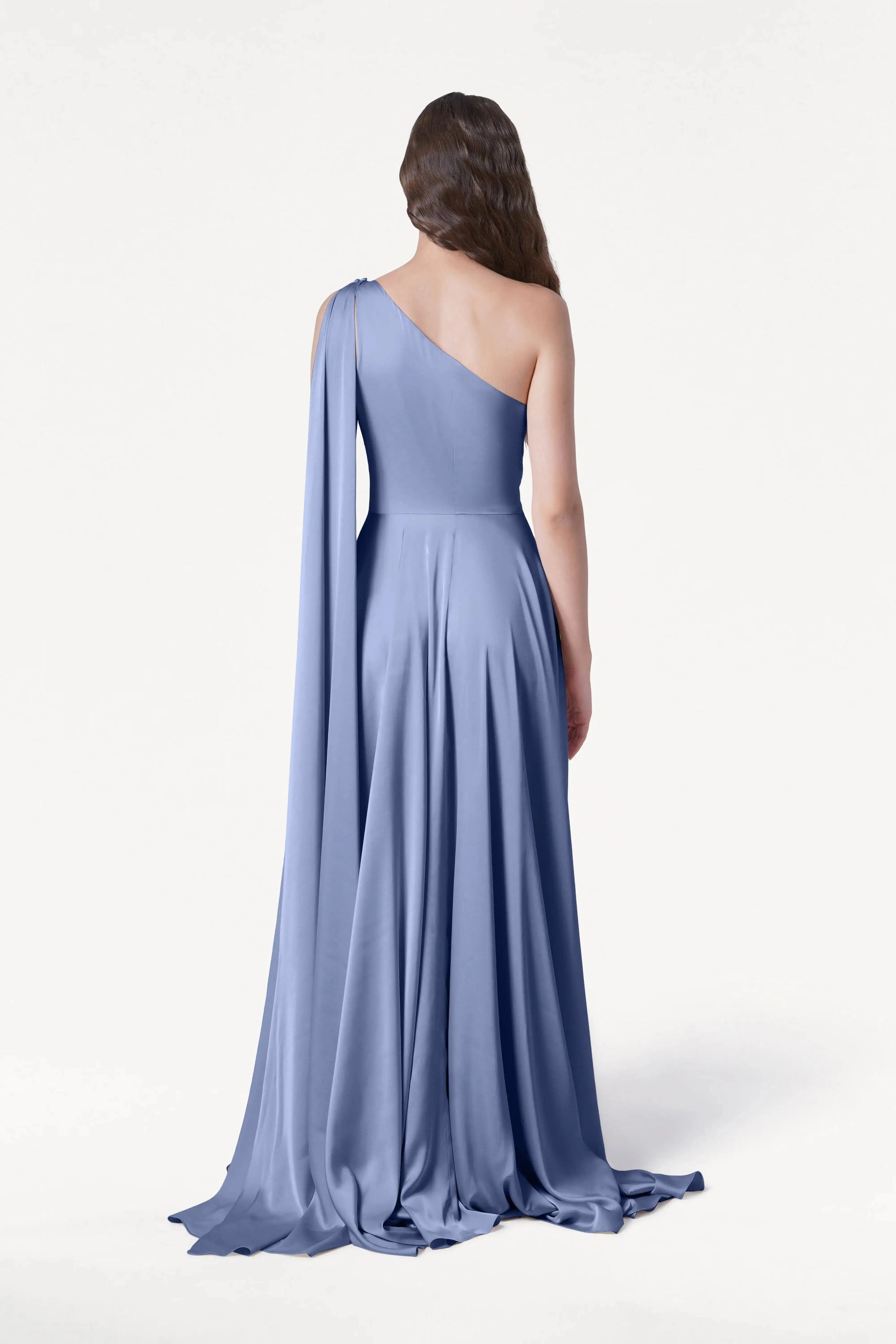 A woman with long wavy hair stands facing away, wearing the TH&TH Lyra Bridesmaid Dress in French Blue Satin—a size-inclusive, floor-length one-shoulder gown with a flowing skirt and draped fabric—against a plain white background.
