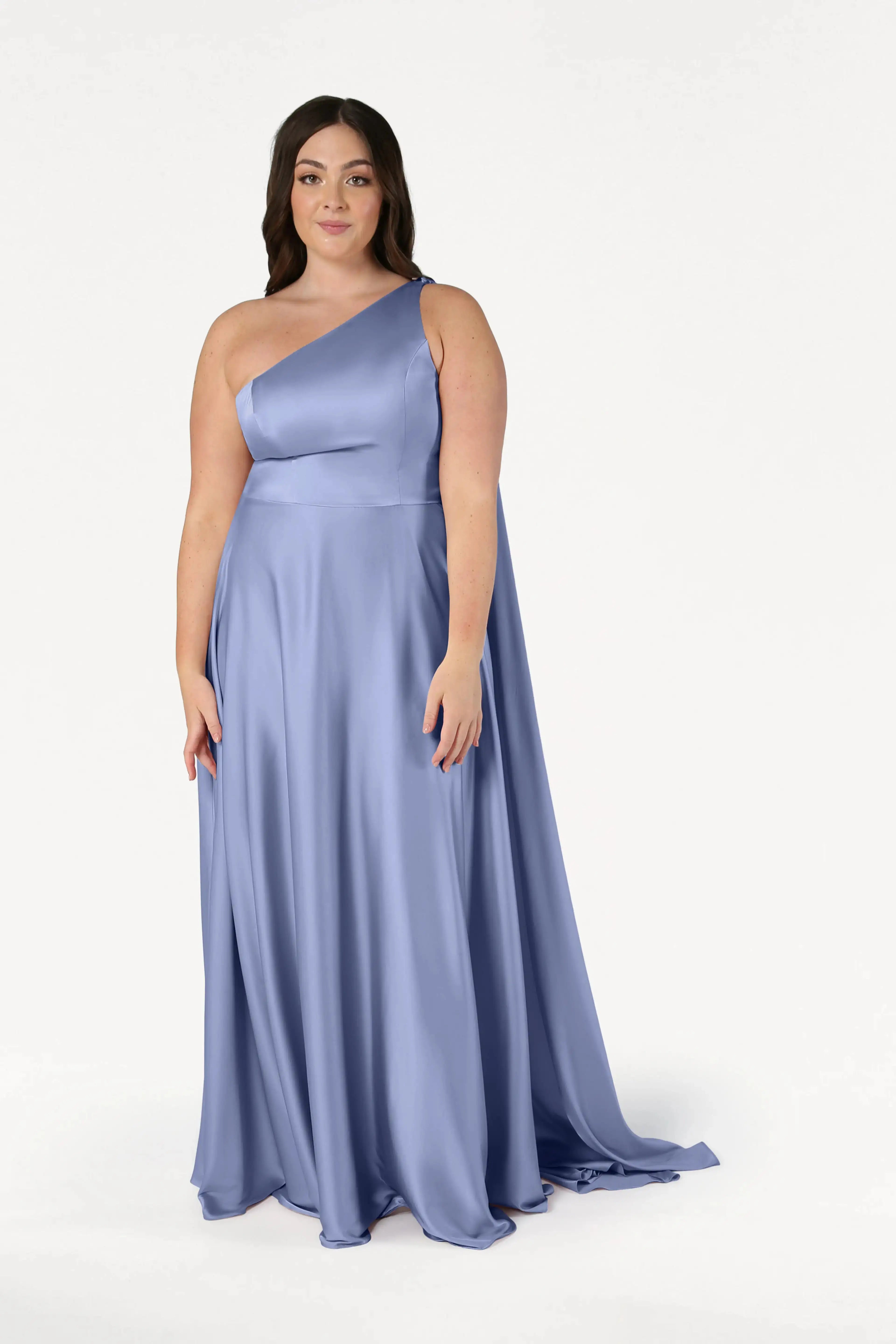 A woman with long dark hair wears the TH&TH Lyra Bridesmaid Dress in French Blue Satin, a flowing, size-inclusive one-shoulder gown with a fitted waist. She faces forward against a white background, smiling softly.