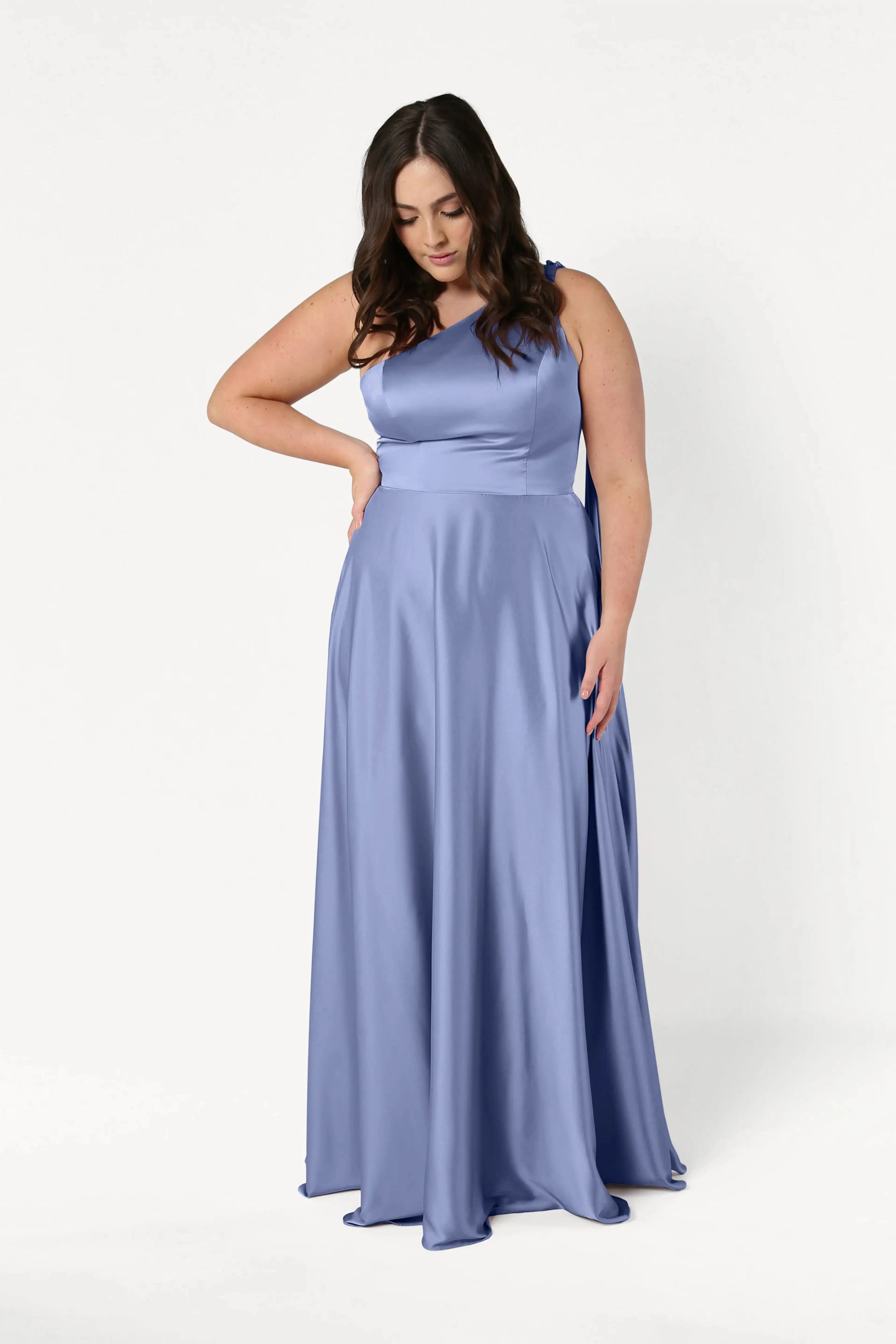 A woman with long dark hair wears the TH&TH Lyra Bridesmaid Dress in French Blue Satin, a size-inclusive, floor-length gown. She stands against a plain white background, looking down with one hand on her waist.