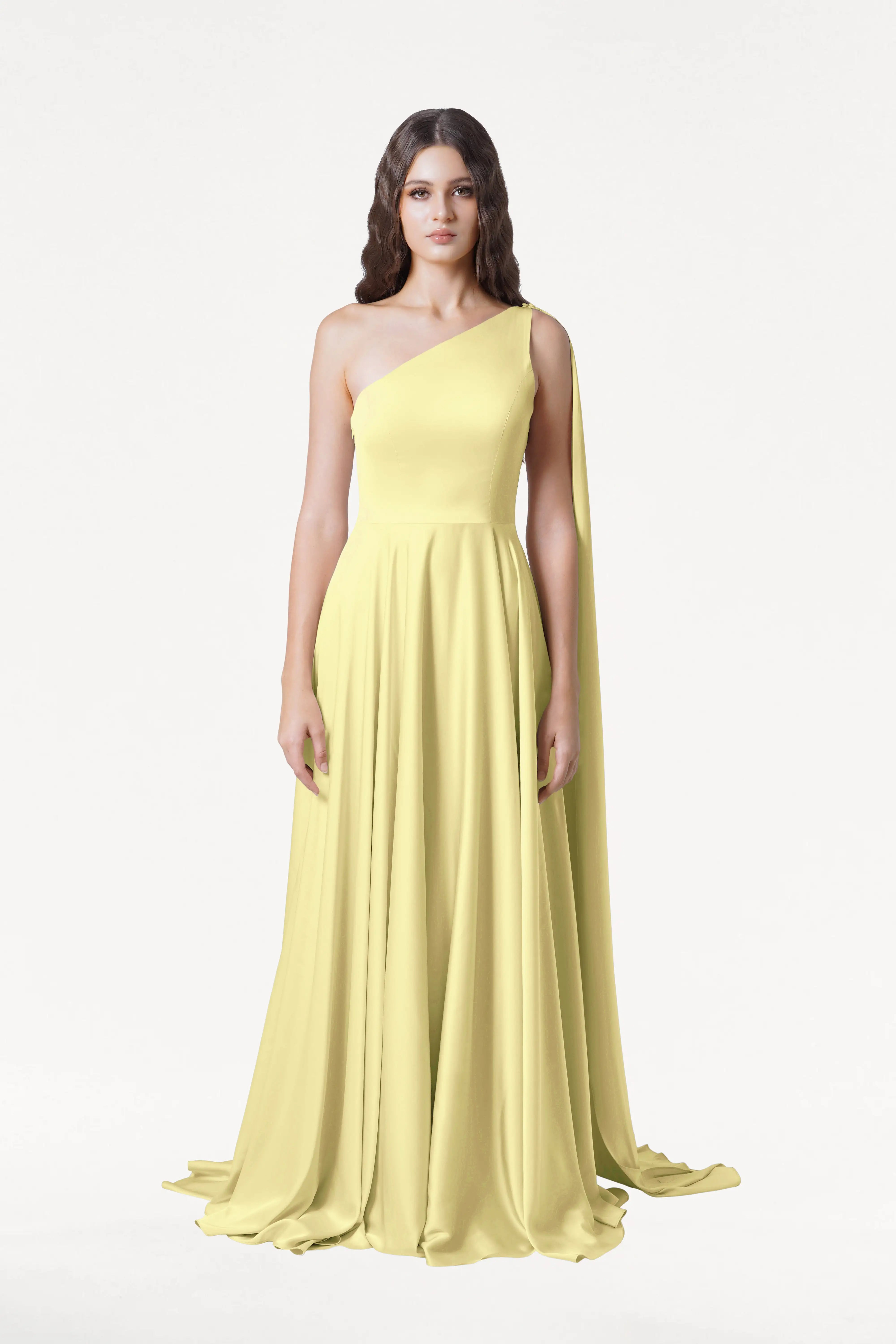 A woman faces forward in the TH&TH Lyra Bridesmaid Dress in Limoncello Satin—a size-inclusive, long, flowing one-shoulder gown with a draped panel. She has long wavy brown hair and a neutral expression against a plain white background.