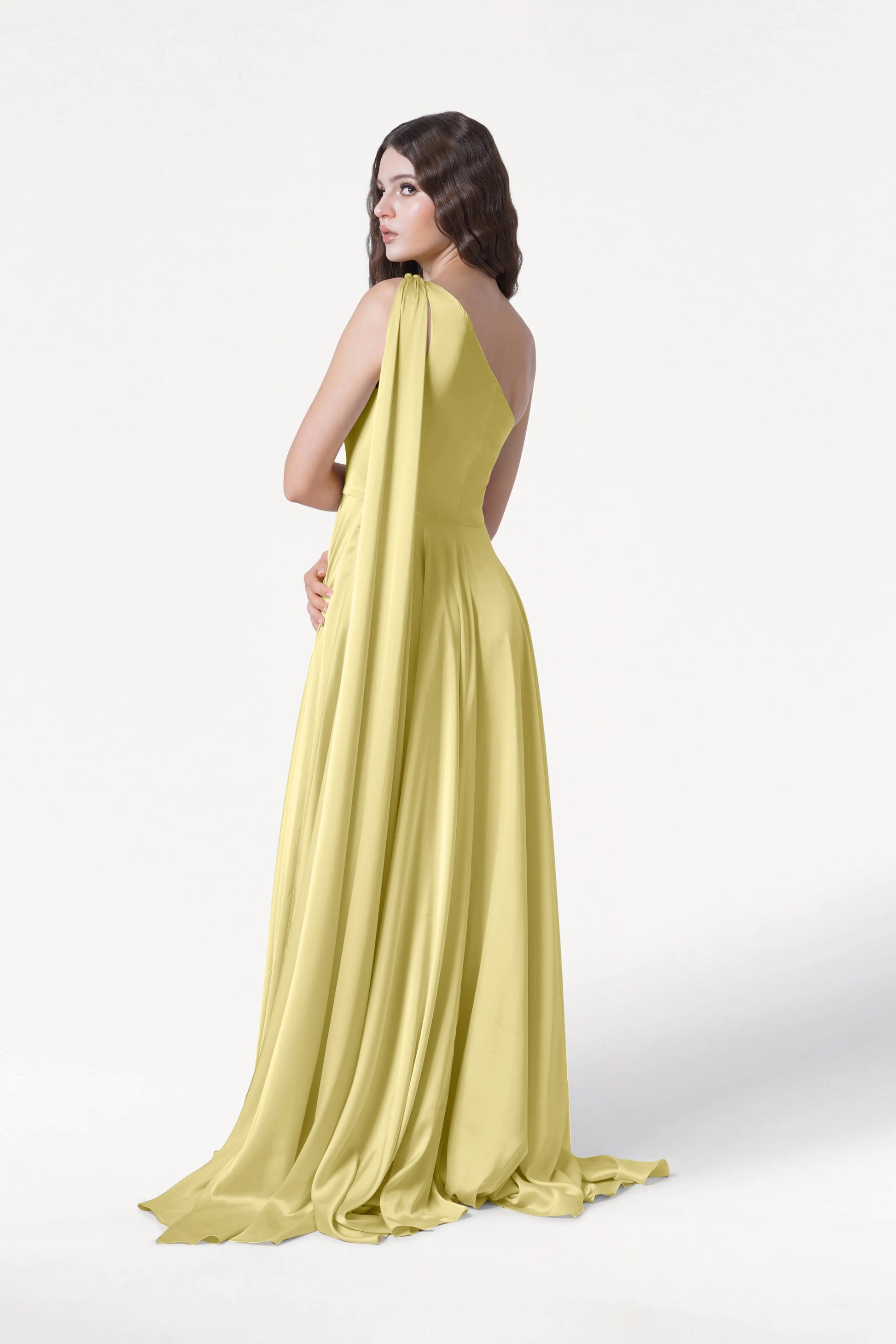 A woman stands against a plain white background, looking over her shoulder in the TH&TH Lyra Bridesmaid Dress in Limoncello Satin—a long, flowing, one-shoulder gown with a draped back.