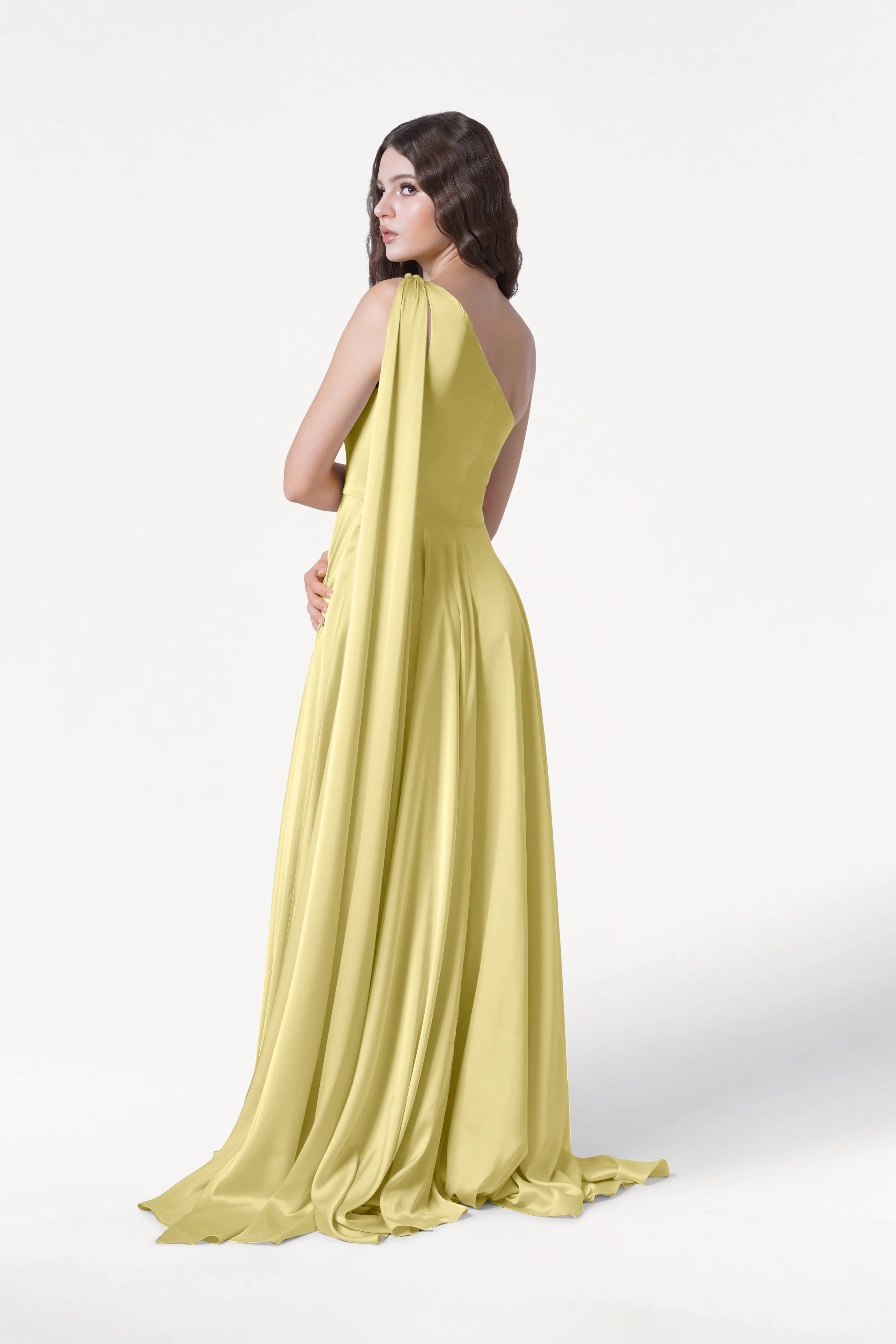A woman stands against a plain white background, looking over her shoulder in the TH&TH Lyra Bridesmaid Dress in Limoncello Satin—a long, flowing, one-shoulder gown with a draped back.