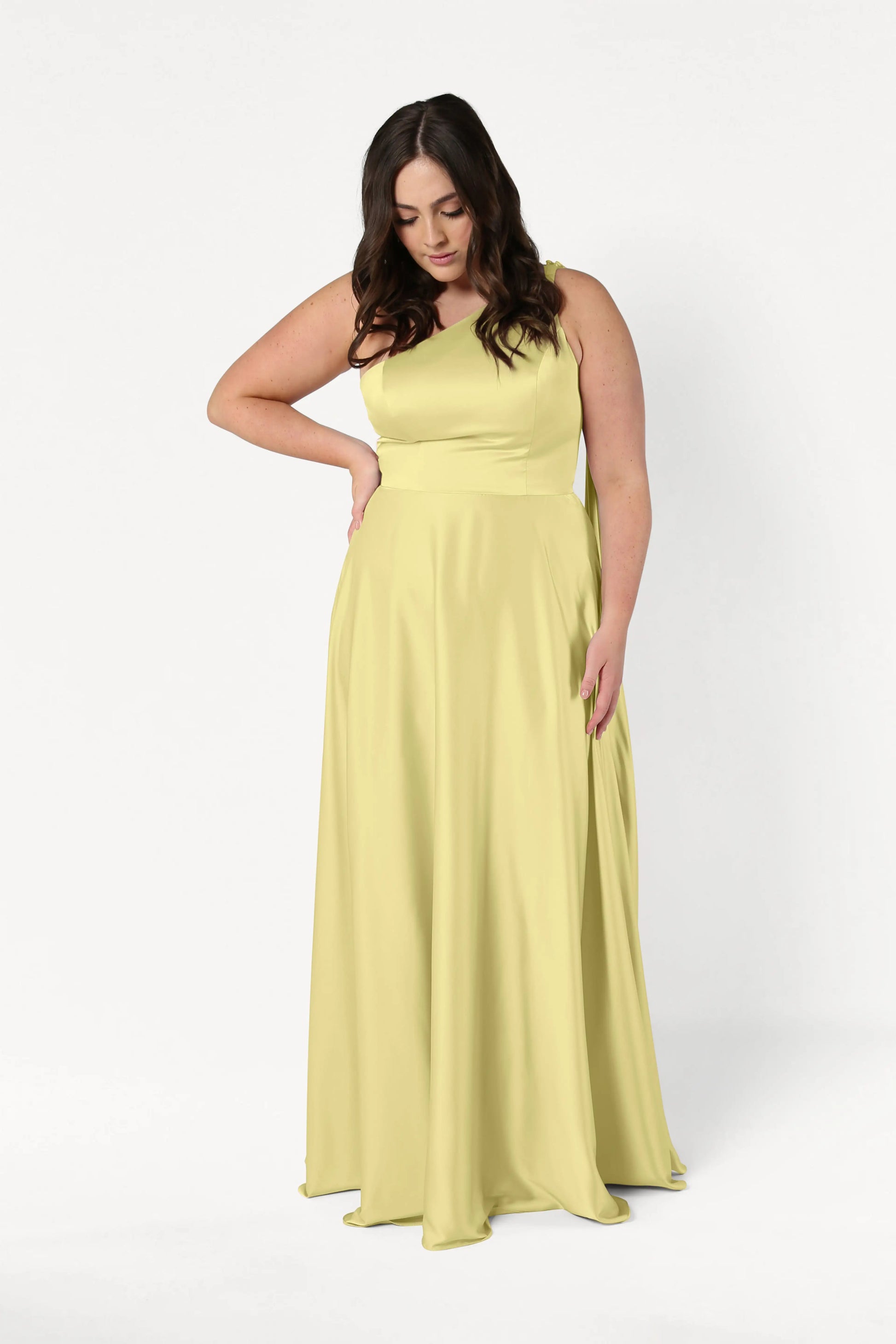 A woman with long dark hair wears the TH&TH Lyra Bridesmaid Dress in Limoncello Satin—a size-inclusive, floor-length gown—as she stands against a plain white background, looking down with one hand on her hip.