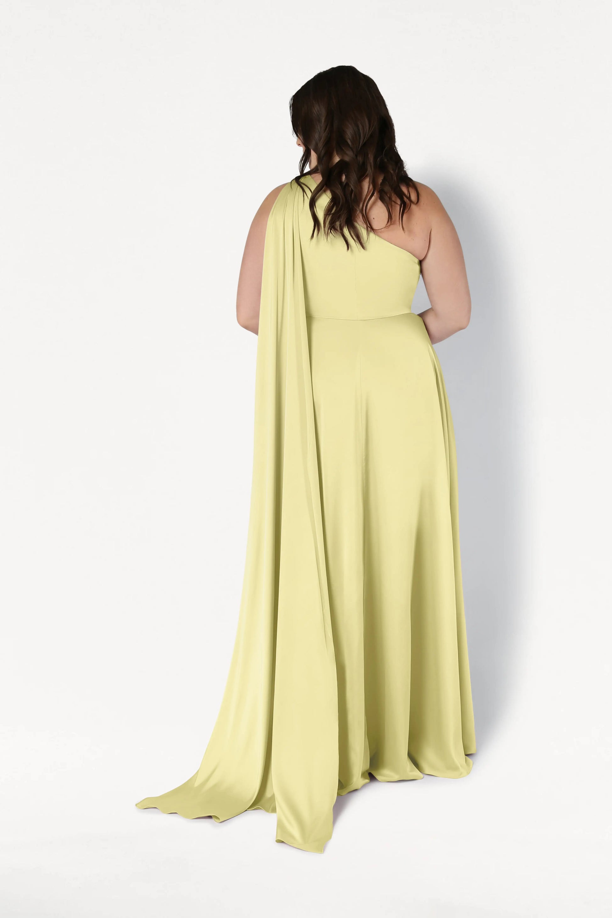 A woman with wavy brown hair stands with her back to the camera against a white background, wearing the size-inclusive Lyra Bridesmaid Dress in Limoncello Satin by TH&TH, featuring a one-shoulder design and long flowing cape.