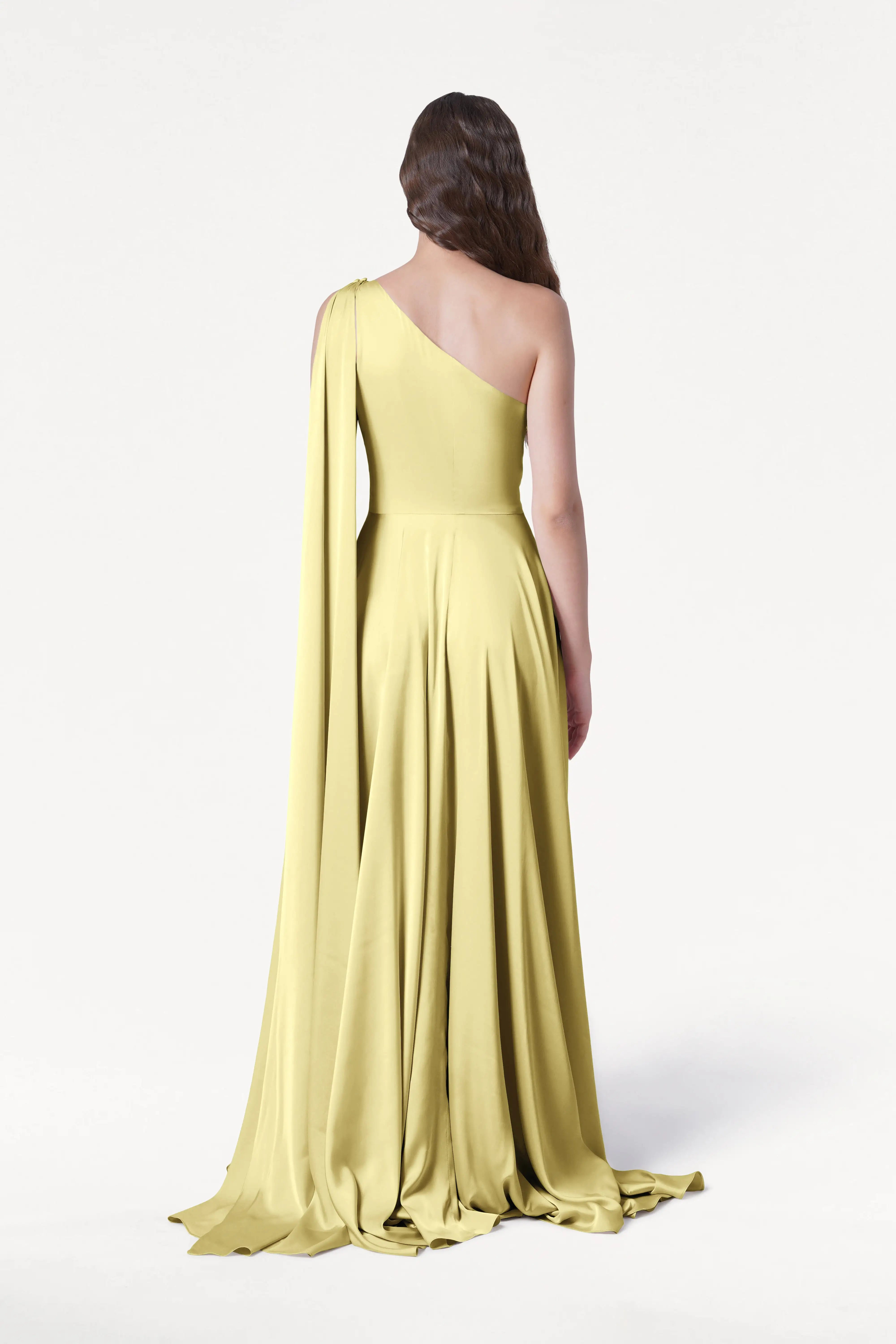 A woman faces away from the camera, modeling the Lyra Bridesmaid Dress in Limoncello Satin by TH&TH—a size-inclusive, pale yellow one-shoulder gown with a draped sash and floor-length hem against a plain white background.