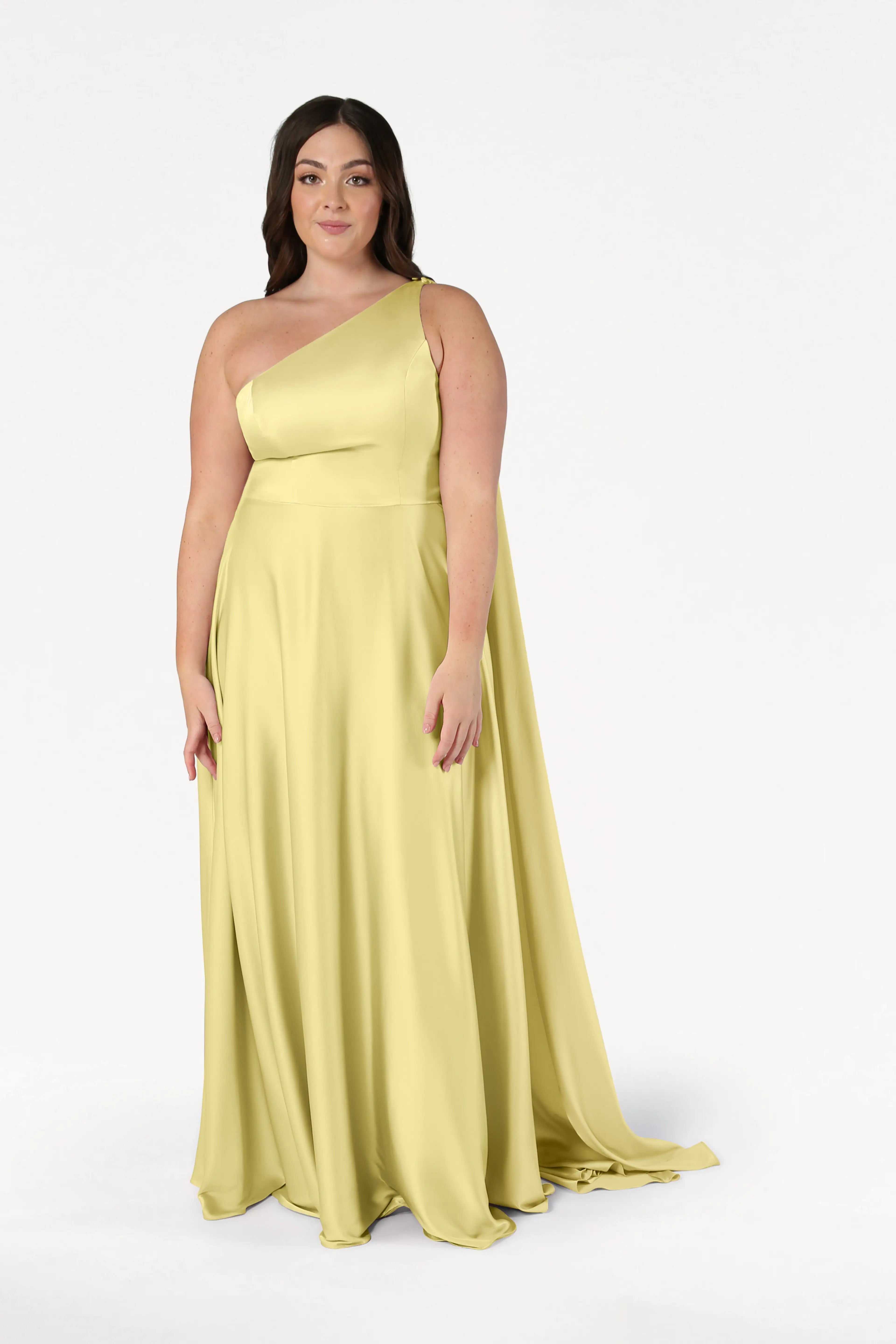 A woman wears the TH&TH Lyra Bridesmaid Dress in Limoncello Satin—a size-inclusive, long one-shoulder gown—standing against a plain white background and looking at the camera with a relaxed expression.