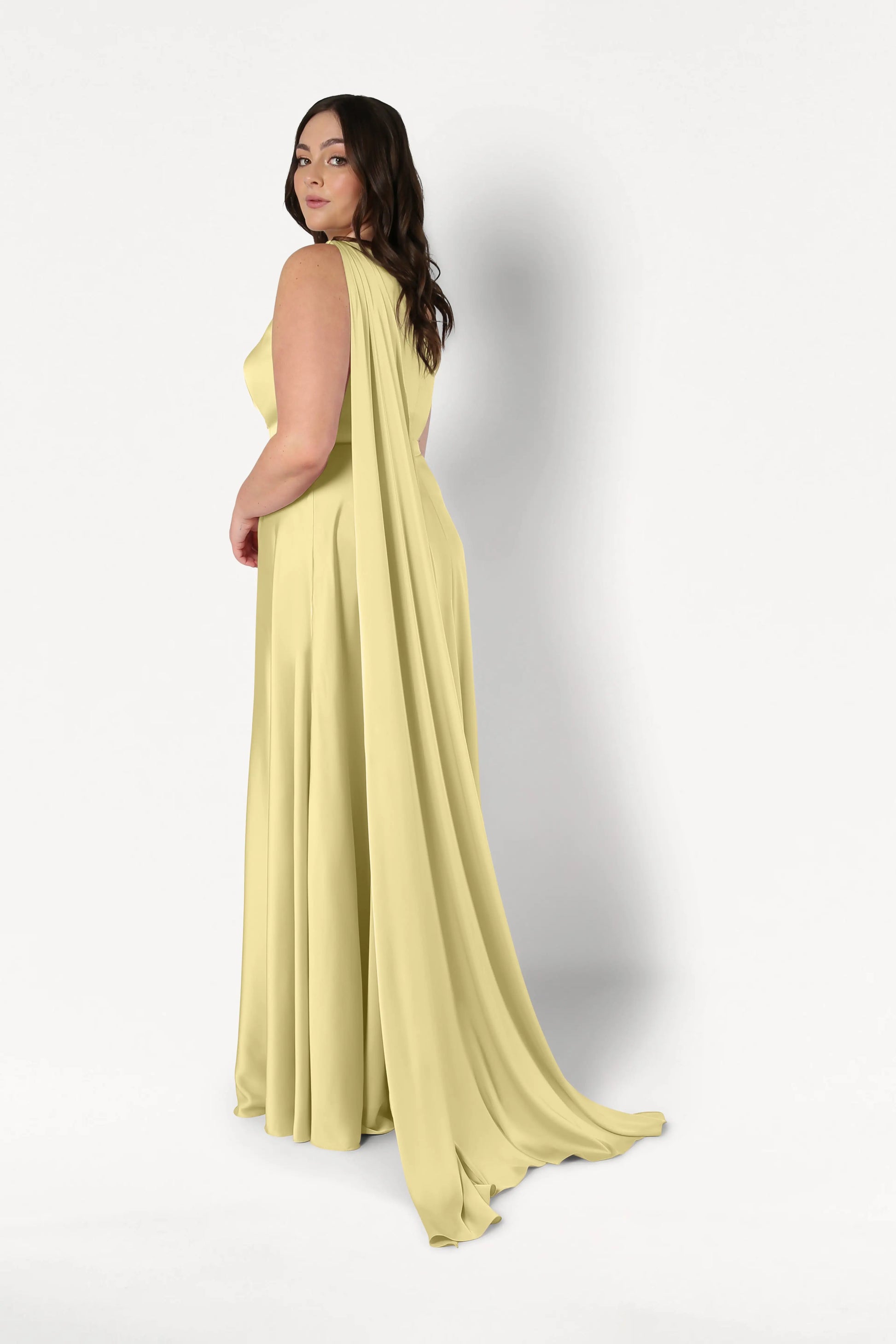 A woman in the Lyra Bridesmaid Dress in Limoncello Satin by TH&TH, featuring a flowing cape and size-inclusive fit, looks over her shoulder against a plain white background.