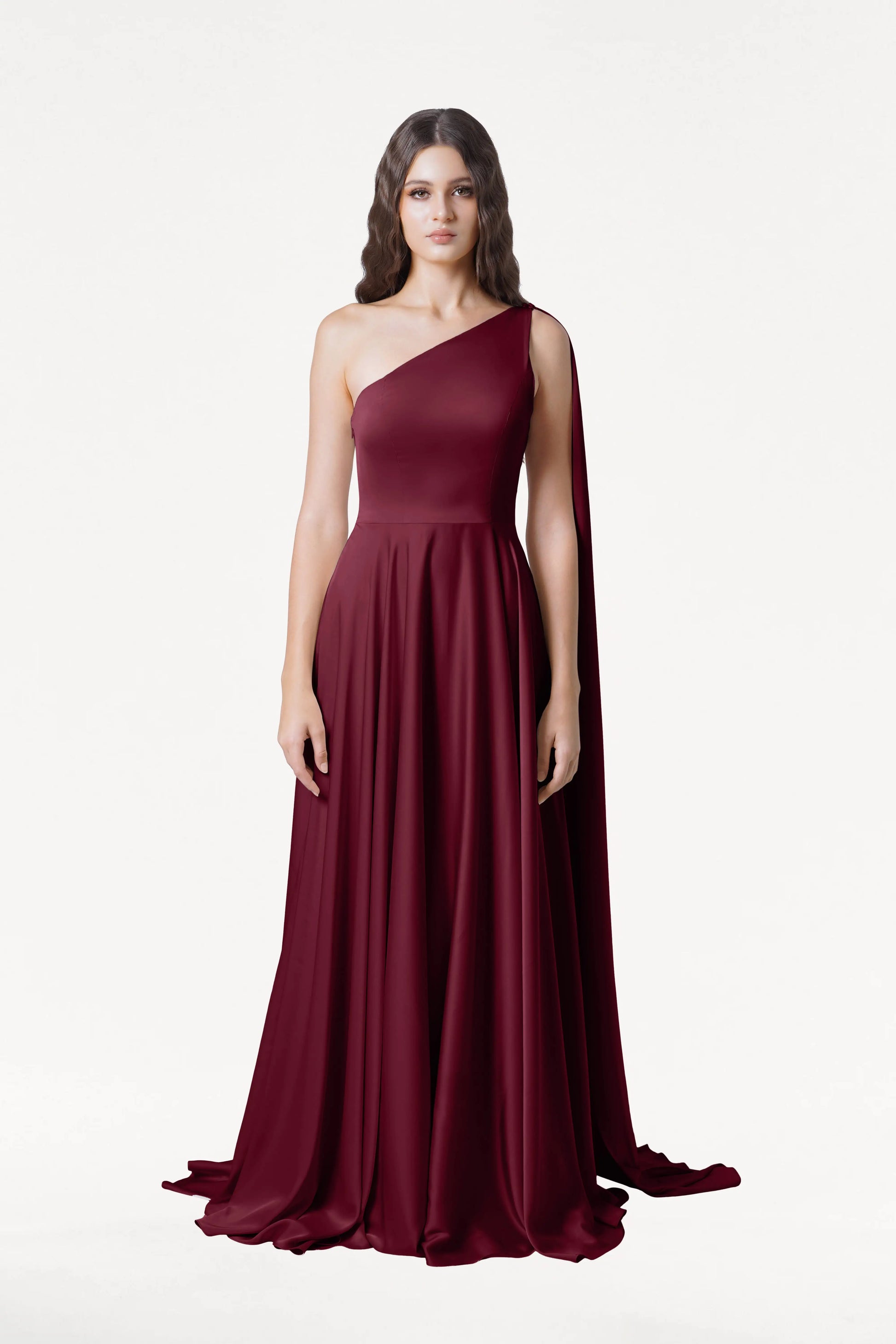 A woman faces forward in the Lyra Bridesmaid Dress in Merlot Satin by TH&TH, a size-inclusive, floor-length one-shoulder gown with a flowing cape detail. She has long wavy hair and a neutral expression against a plain white background.