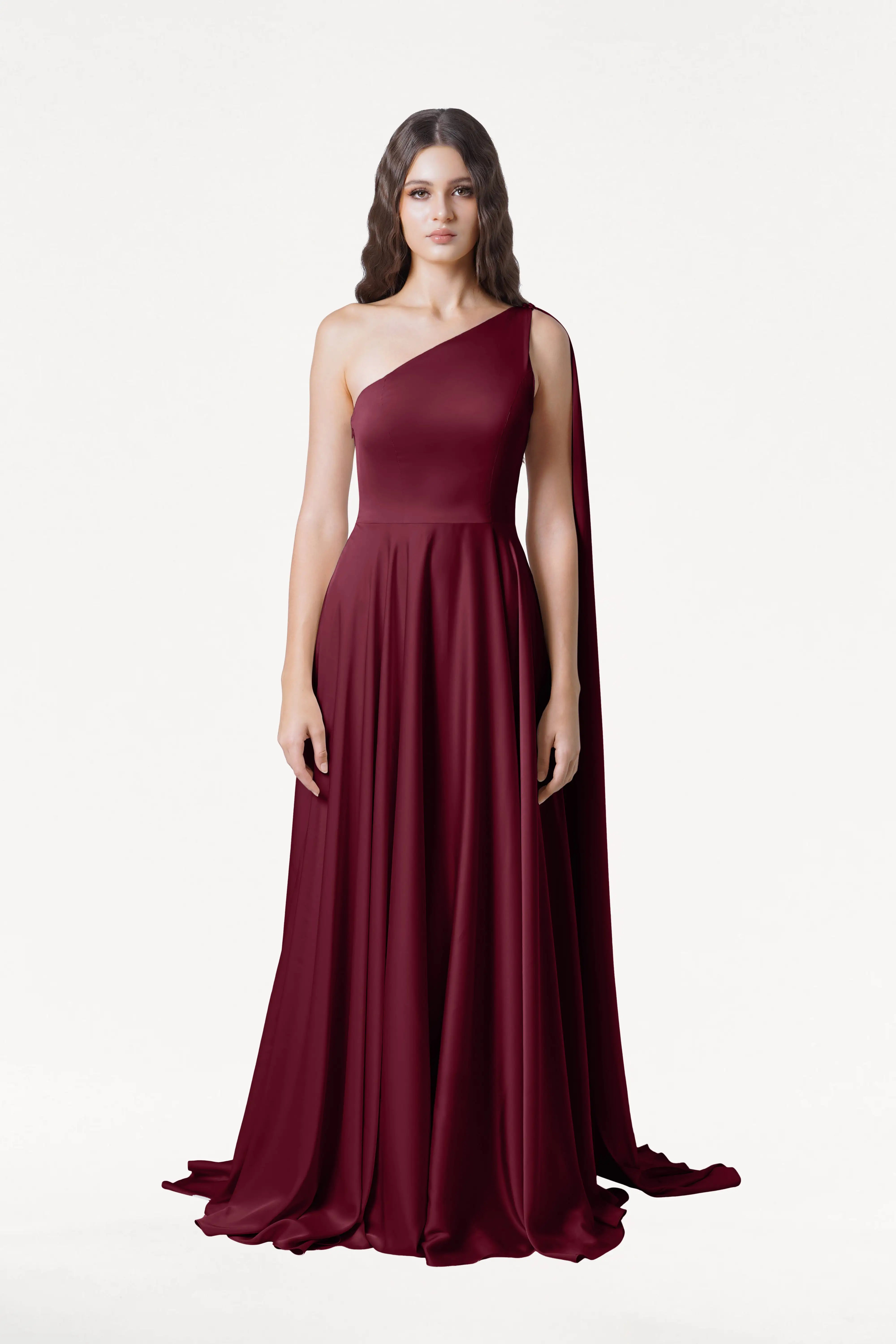 A woman faces forward in the Lyra Bridesmaid Dress in Merlot Satin by TH&TH, a size-inclusive, floor-length one-shoulder gown with a flowing cape detail. She has long wavy hair and a neutral expression against a plain white background.