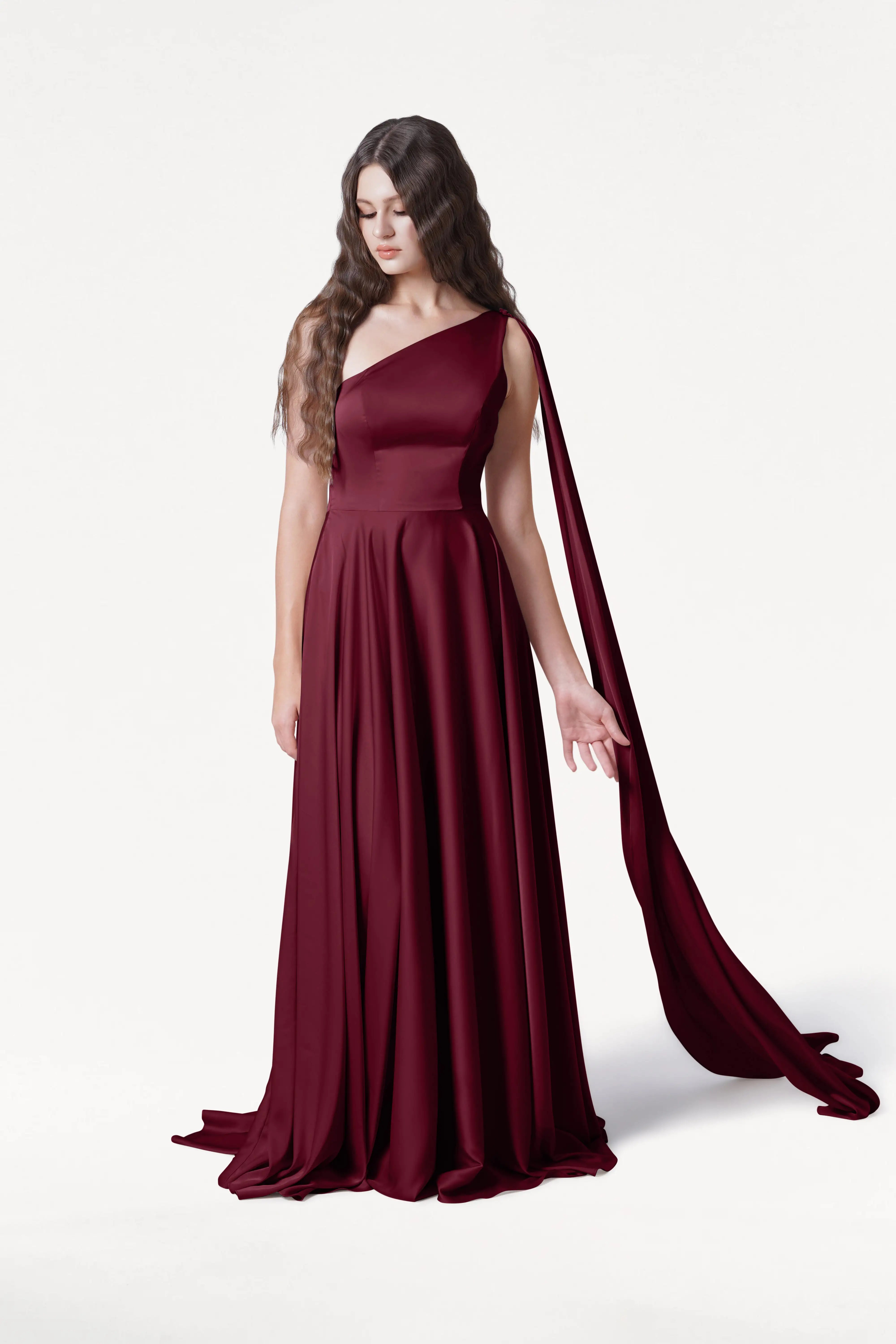 A woman with long wavy brown hair wears the TH&TH Lyra Bridesmaid Dress in Merlot Satin, a size-inclusive, floor-length one-shoulder gown with a flowing sash, standing against a plain white background.