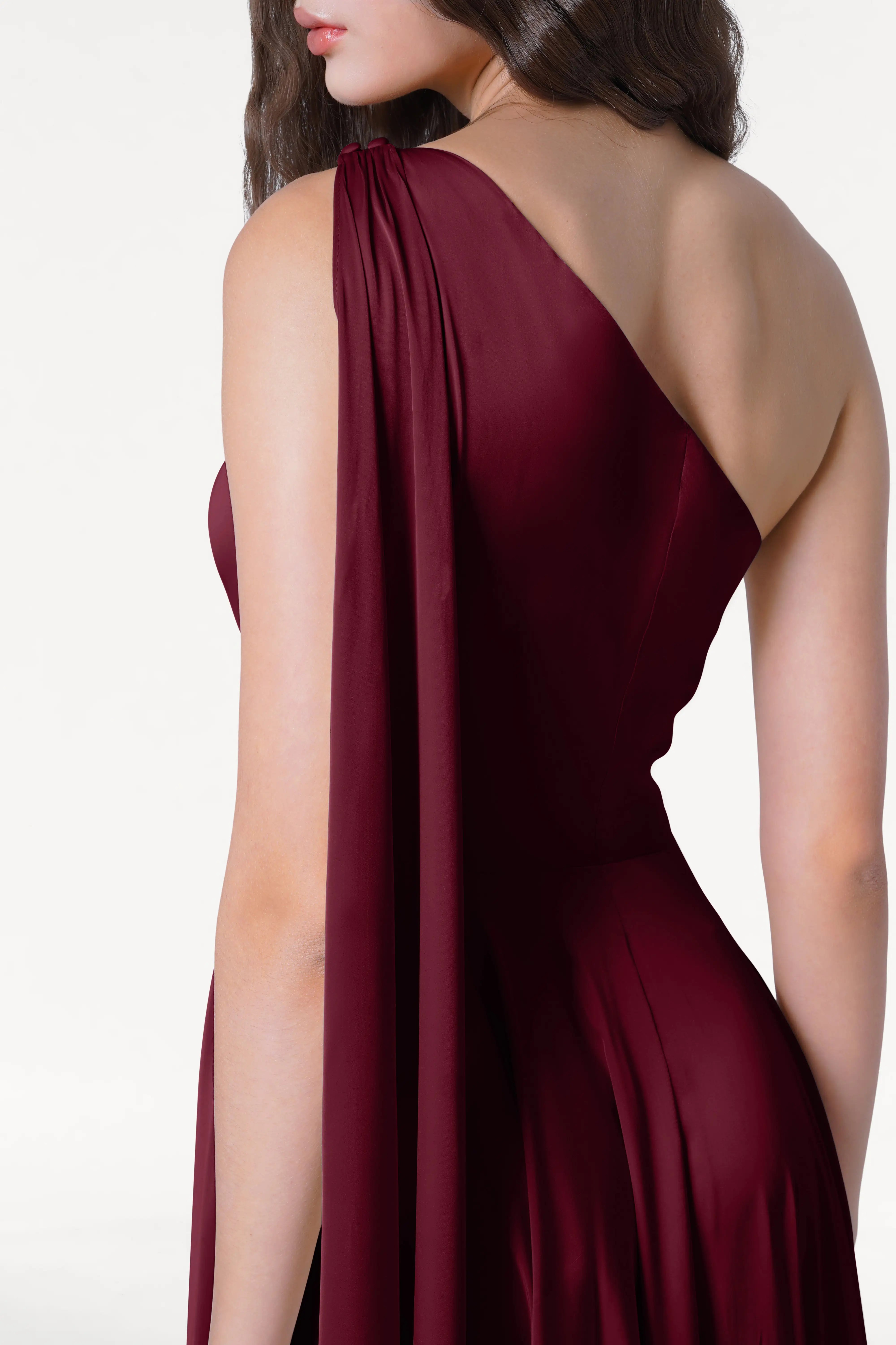 A woman is shown from the back and side in the TH&TH Lyra Bridesmaid Dress in Merlot Satin, a size-inclusive, sleeveless, one-shoulder gown with draped fabric detail. Her long, wavy hair falls down her back against a plain white background.