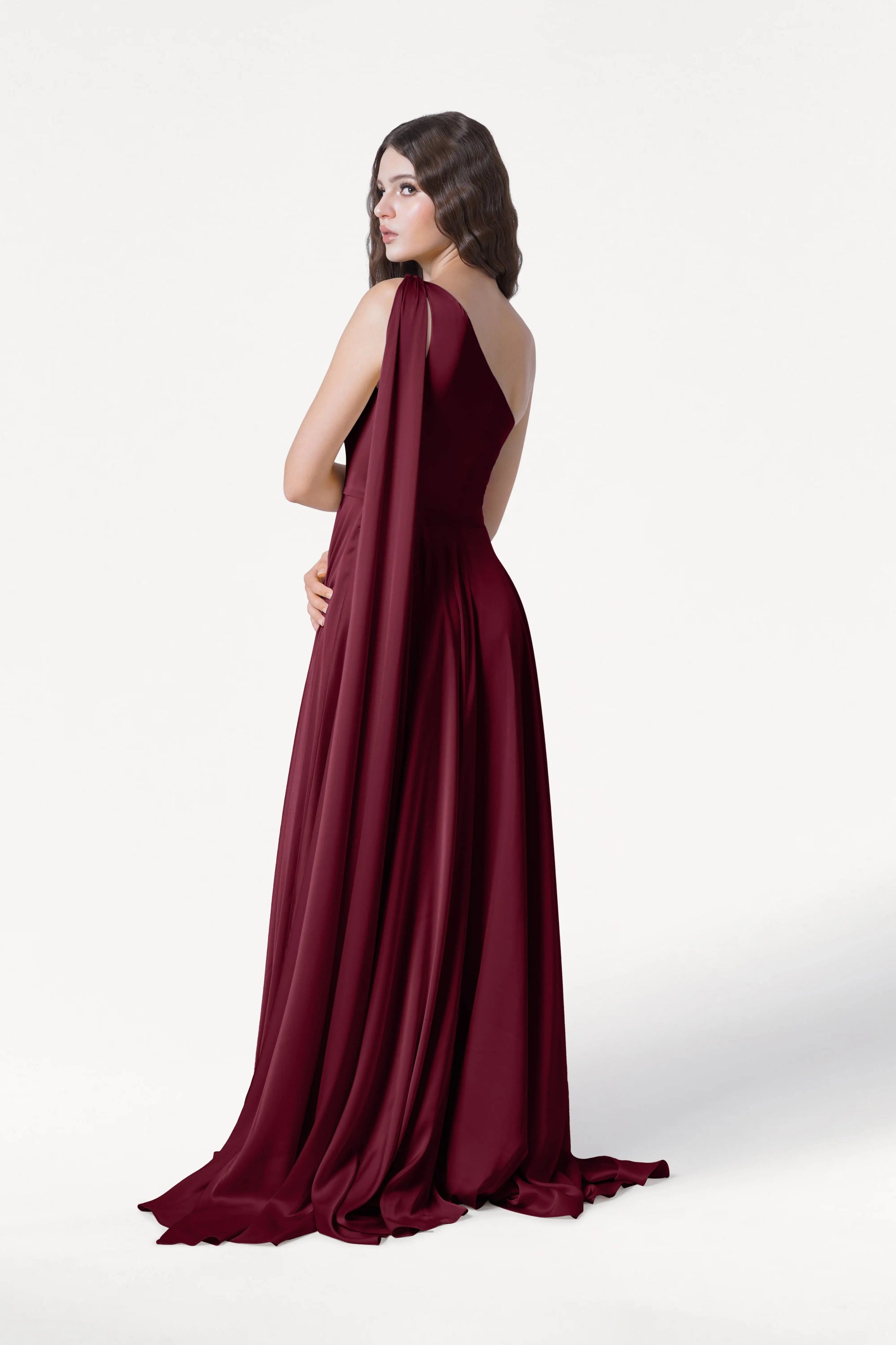 A woman with long, wavy brown hair models the Lyra Bridesmaid Dress in Merlot Satin by TH&TH—an elegant, size-inclusive satin gown with a draped shoulder detail and floor-length silhouette—against a plain white background.