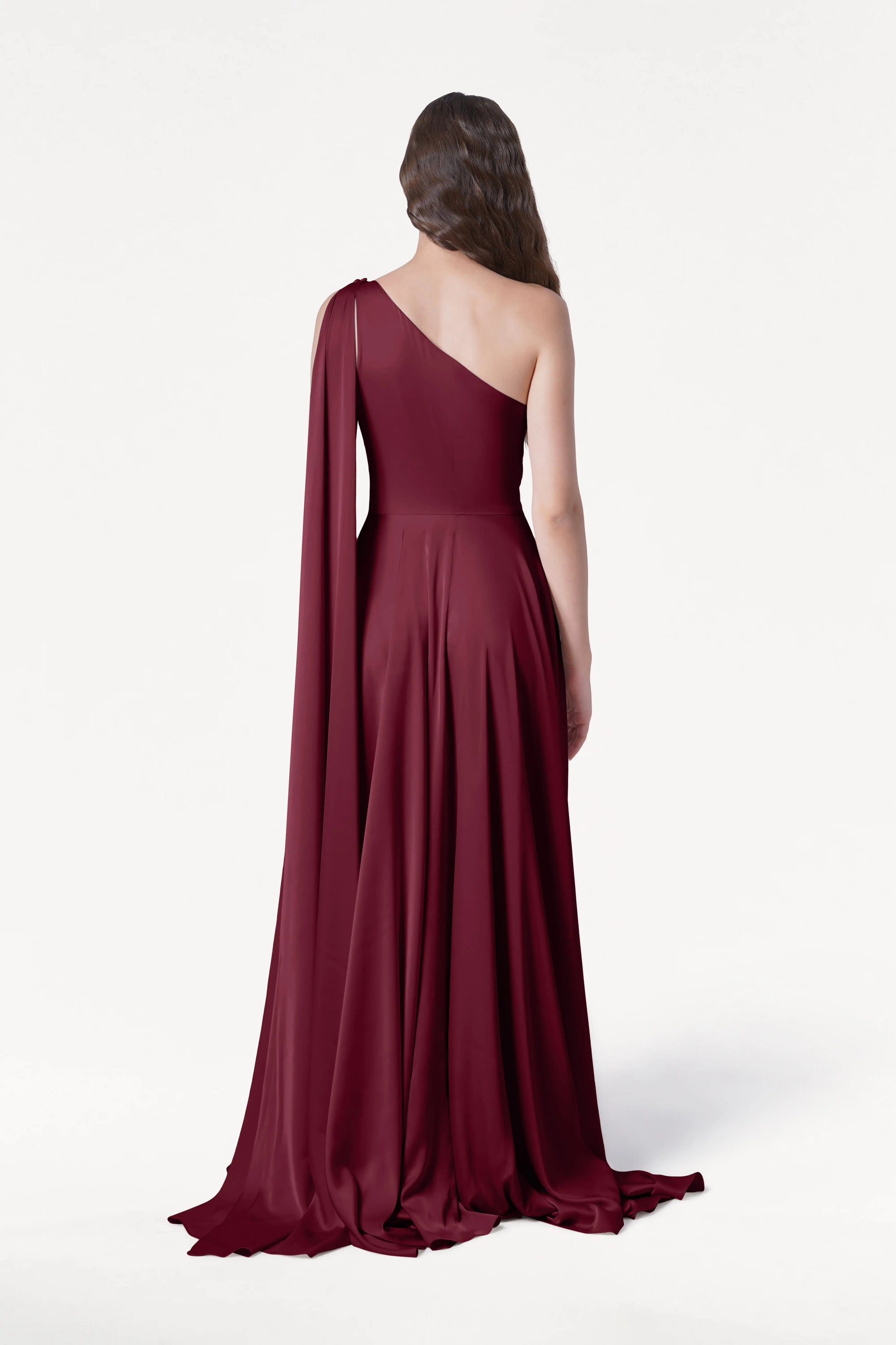 A woman with long, wavy brown hair faces away in the TH&TH Lyra Bridesmaid Dress in Merlot Satin—a size-inclusive, floor-length one-shoulder gown with a draped train—shown against a plain white background.