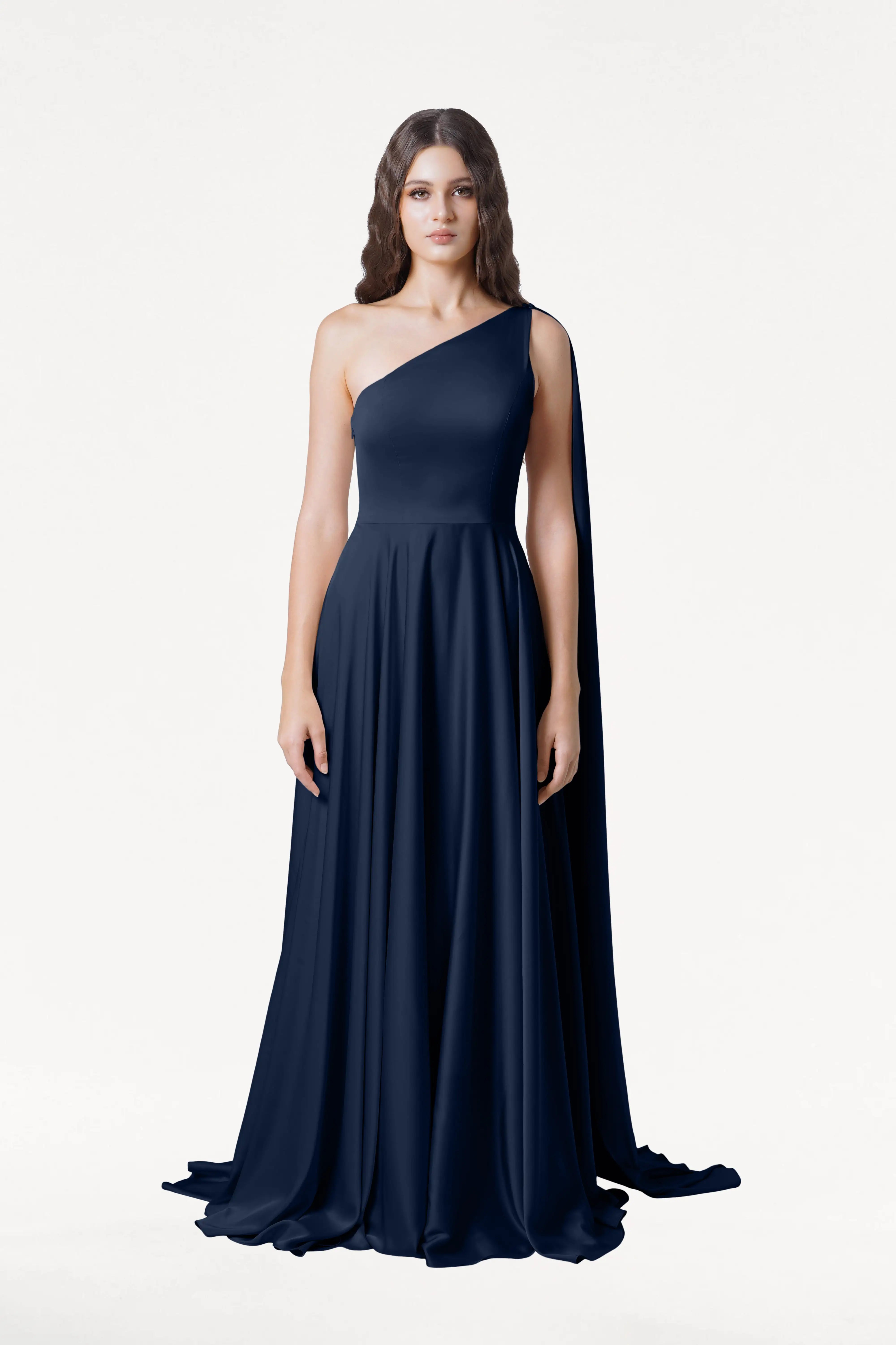 A woman with long wavy brown hair wears the Lyra Bridesmaid Dress in Navy Satin by TH&TH, featuring a floor-length, size-inclusive one-shoulder design. She stands against a plain white background, looking directly at the camera.