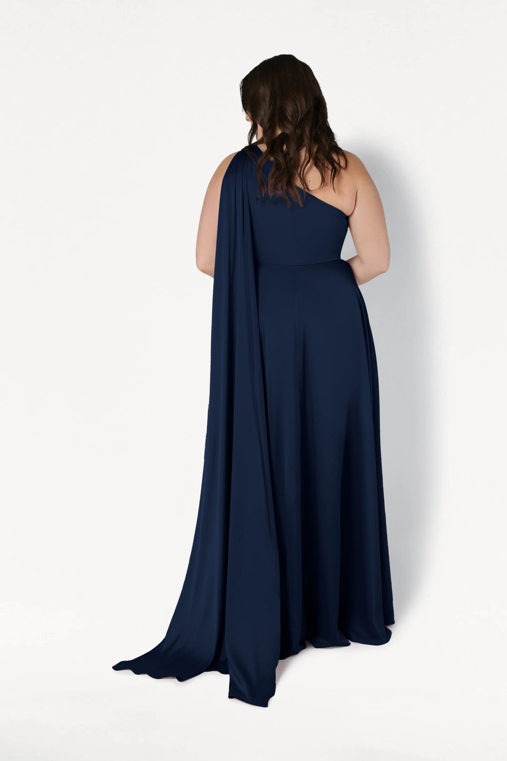 A woman with wavy brown hair faces away, wearing the TH&TH Lyra Bridesmaid Dress in Navy Satin—a size-inclusive, floor-length, one-shoulder gown with a flowing drape—set against a plain white background.