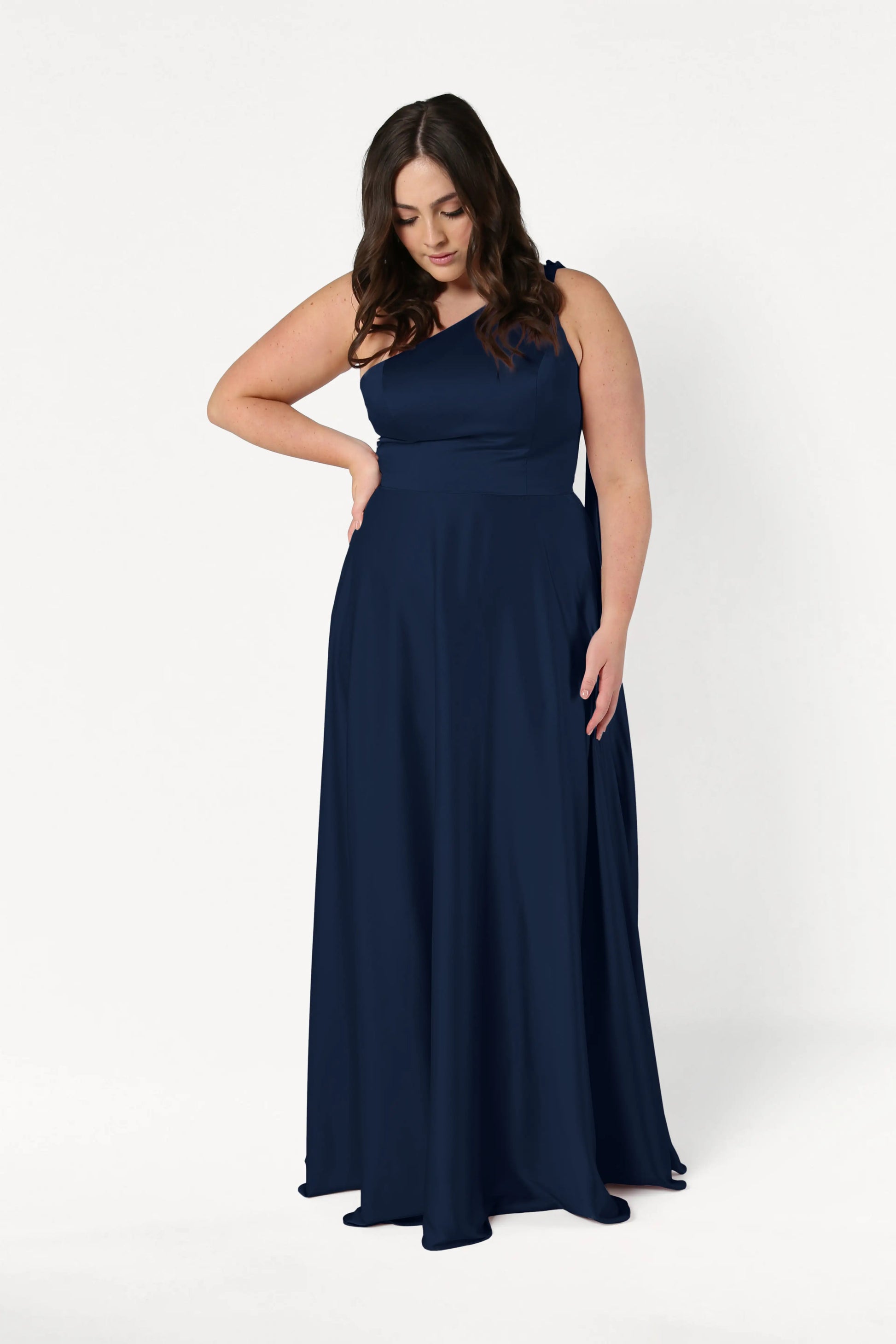 A woman with long brown hair models the TH&TH Lyra Bridesmaid Dress in Navy Satin, standing against a plain white background and looking down with one hand on her waist.