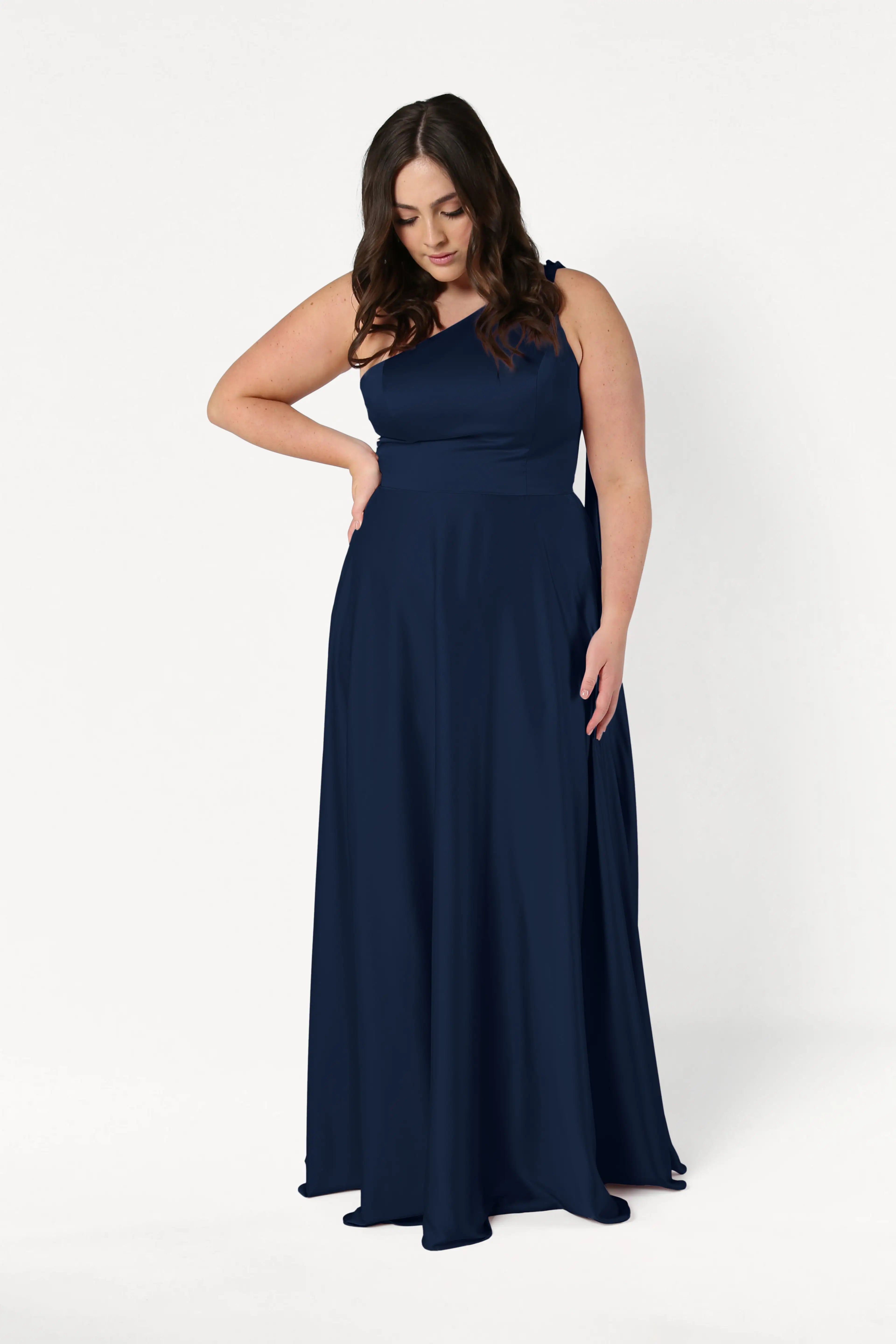 A woman with long brown hair models the TH&TH Lyra Bridesmaid Dress in Navy Satin, standing against a plain white background and looking down with one hand on her waist.