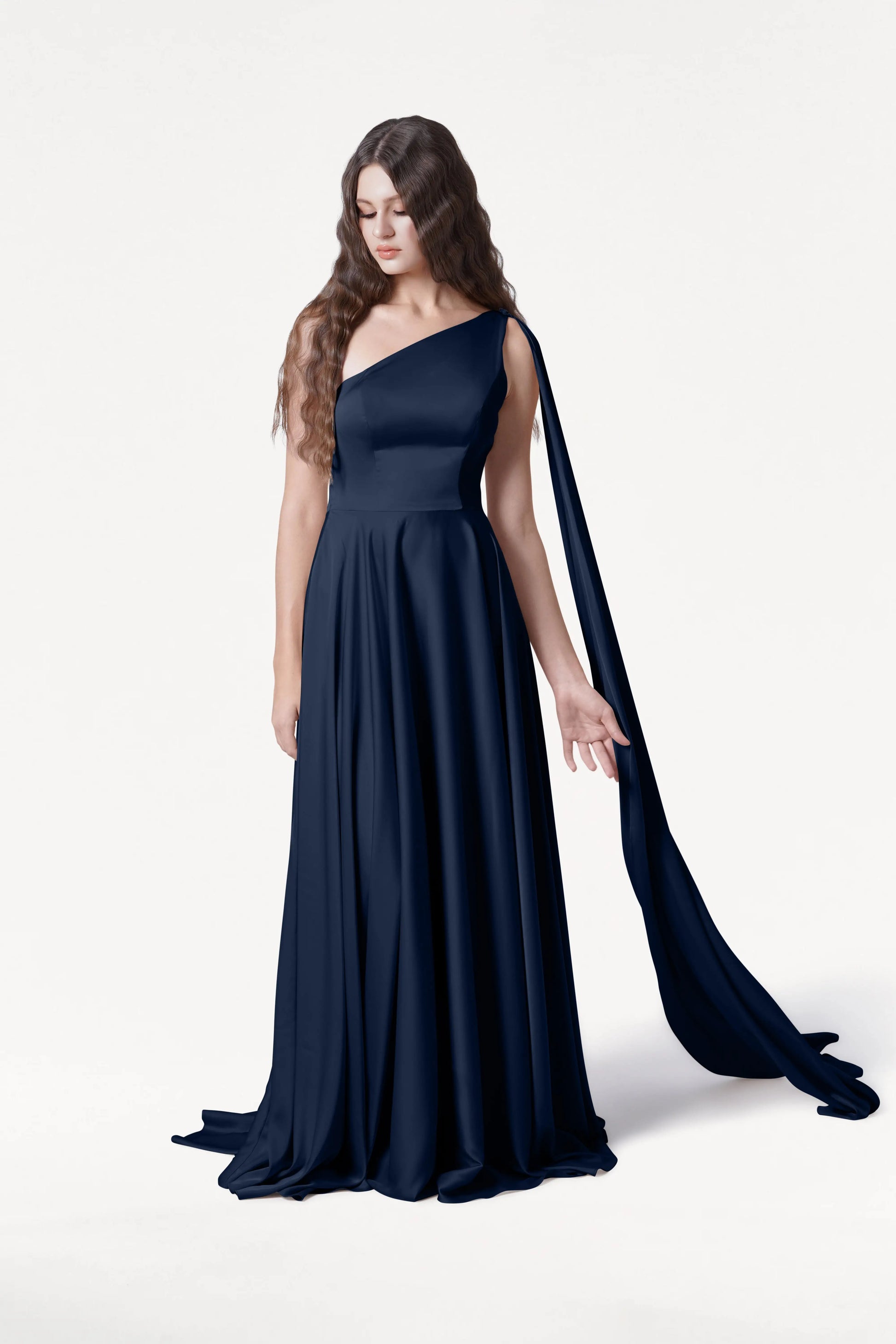 A woman with long, wavy brown hair wears the TH&TH Lyra Bridesmaid Dress in Navy Satin, a size-inclusive, floor-length one-shoulder gown with a flowing drape, standing against a plain white background and looking downwards with one hand extended.