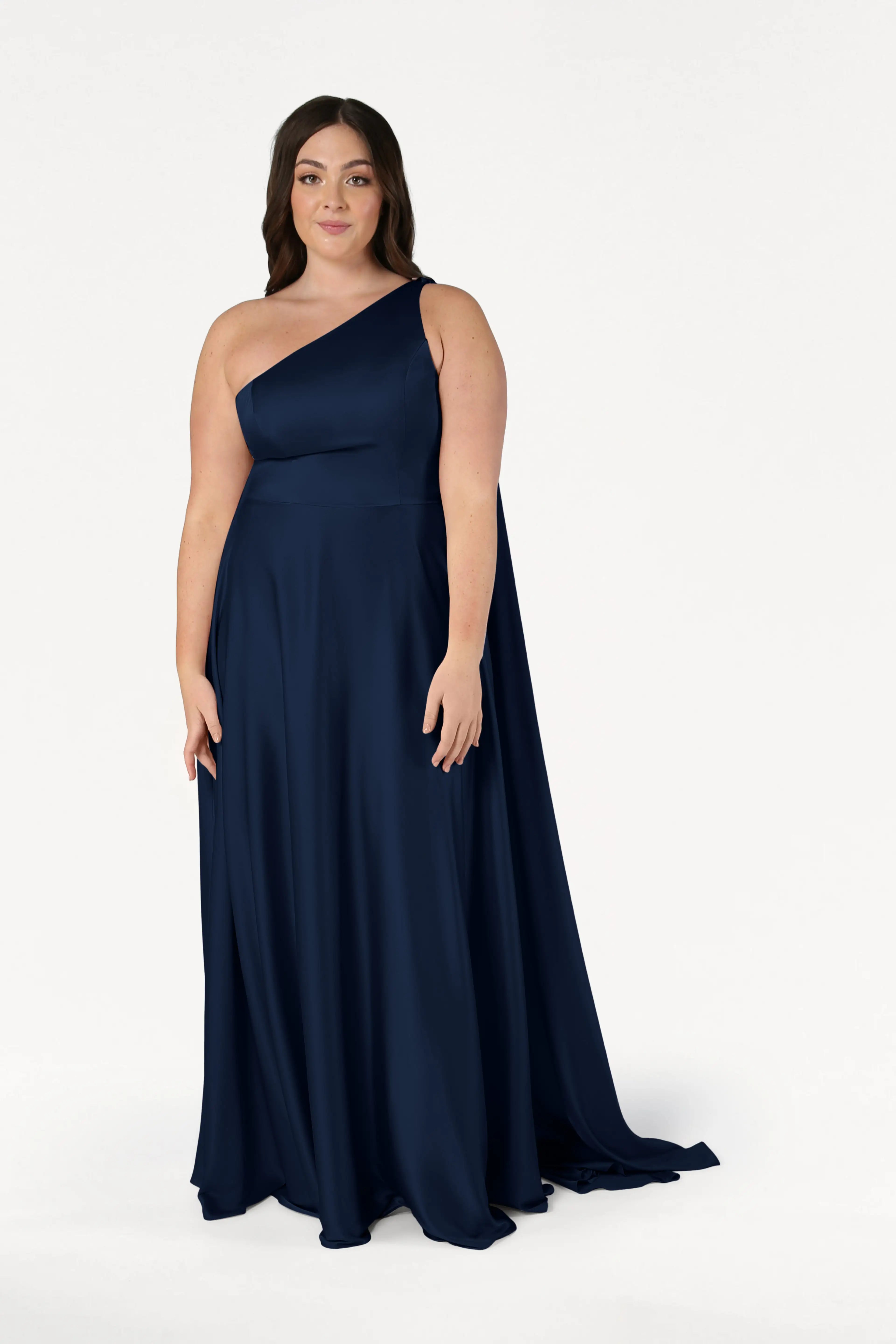 A woman with long brown hair wears the Lyra Bridesmaid Dress in Navy Satin by TH&TH, featuring a size-inclusive fit and a one-shoulder neckline. She stands against a white background, looking at the camera with a slight smile.