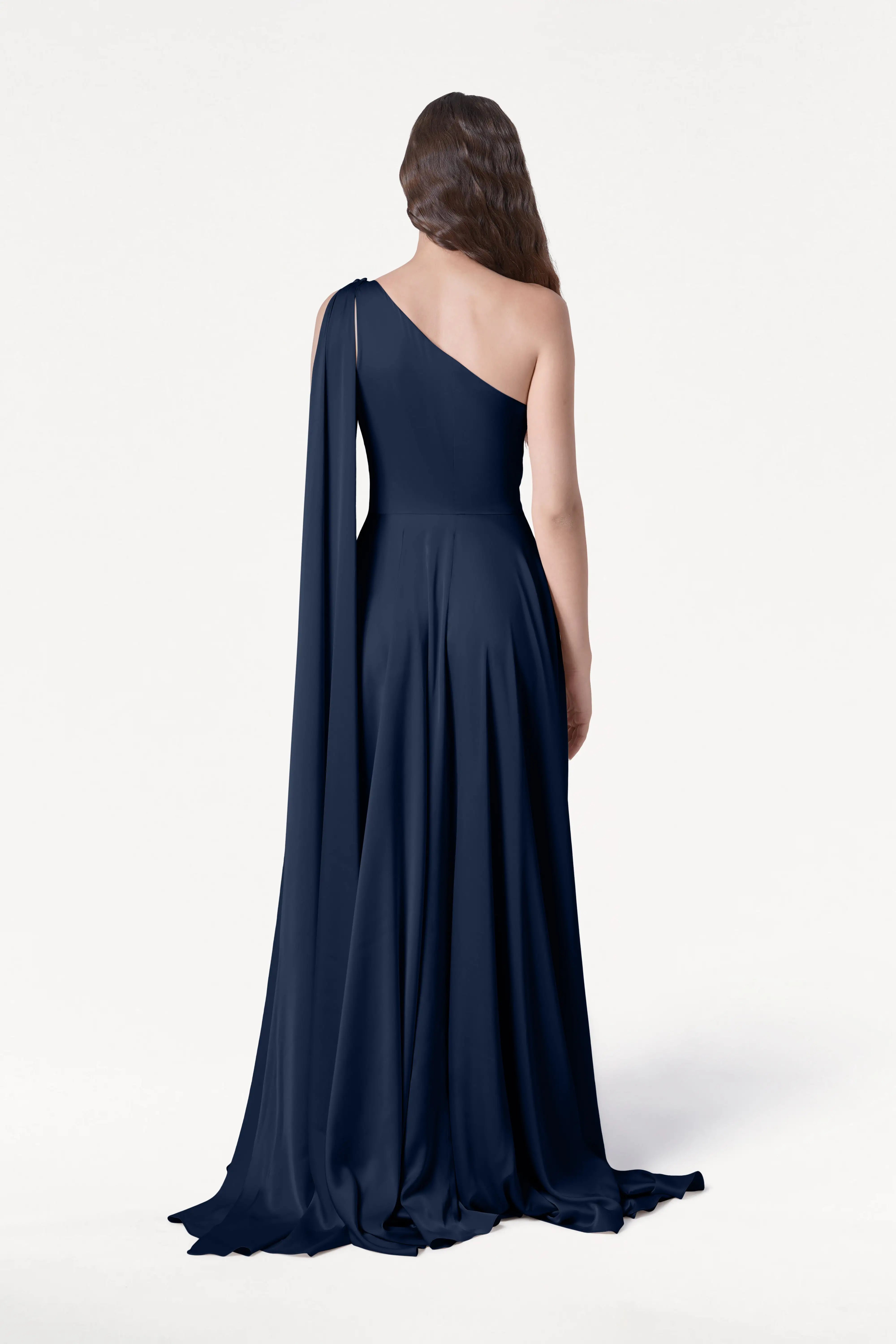 A woman with long brown hair faces away, wearing the TH&TH Lyra Bridesmaid Dress in Navy Satin—a size-inclusive, floor-length one-shoulder gown with a draped train—against a plain white background.