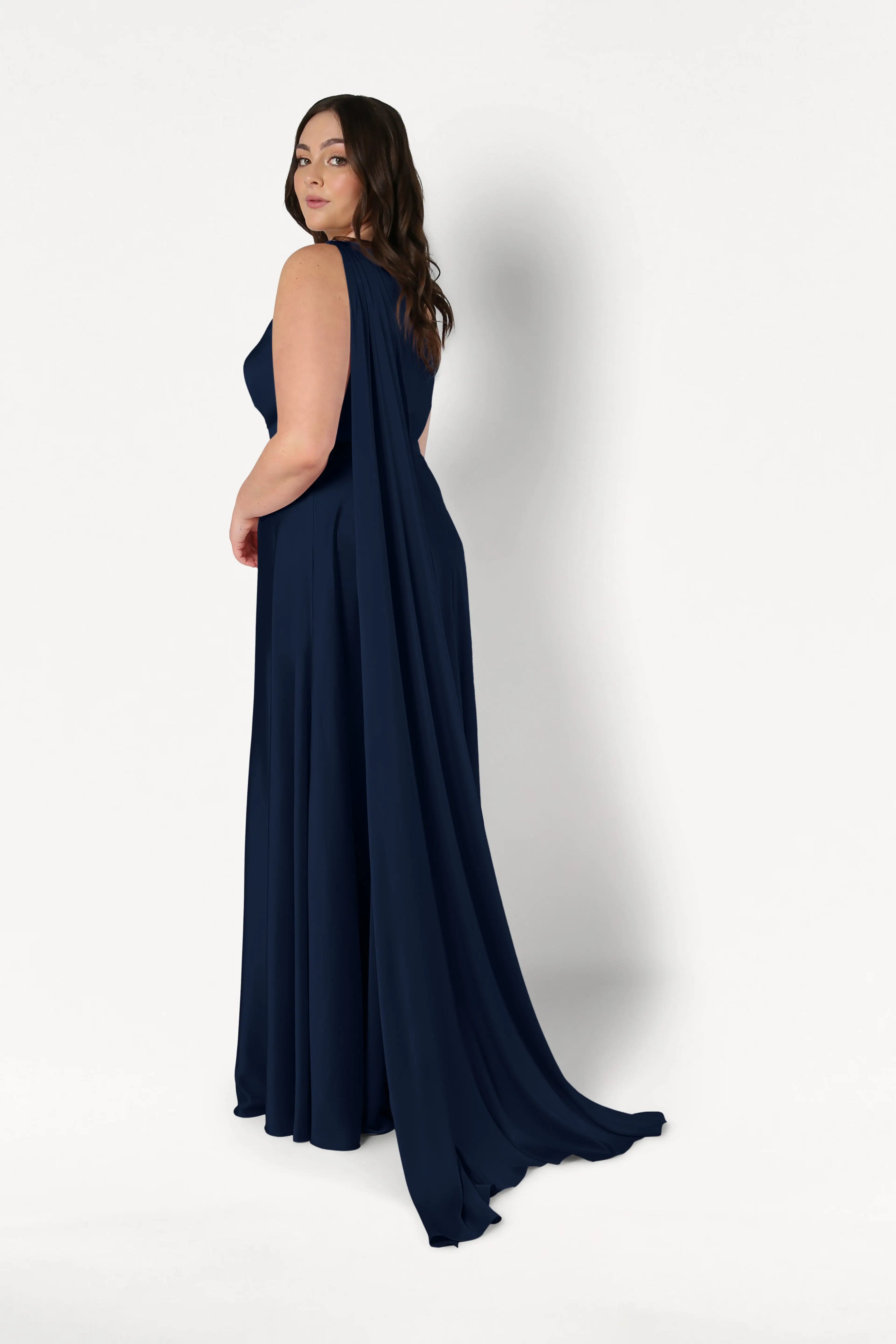 A woman wears the TH&TH Lyra Bridesmaid Dress in Navy Satin, featuring a size-inclusive fit and one-shoulder design with a flowing cape, as she stands against a plain white background looking over her shoulder.