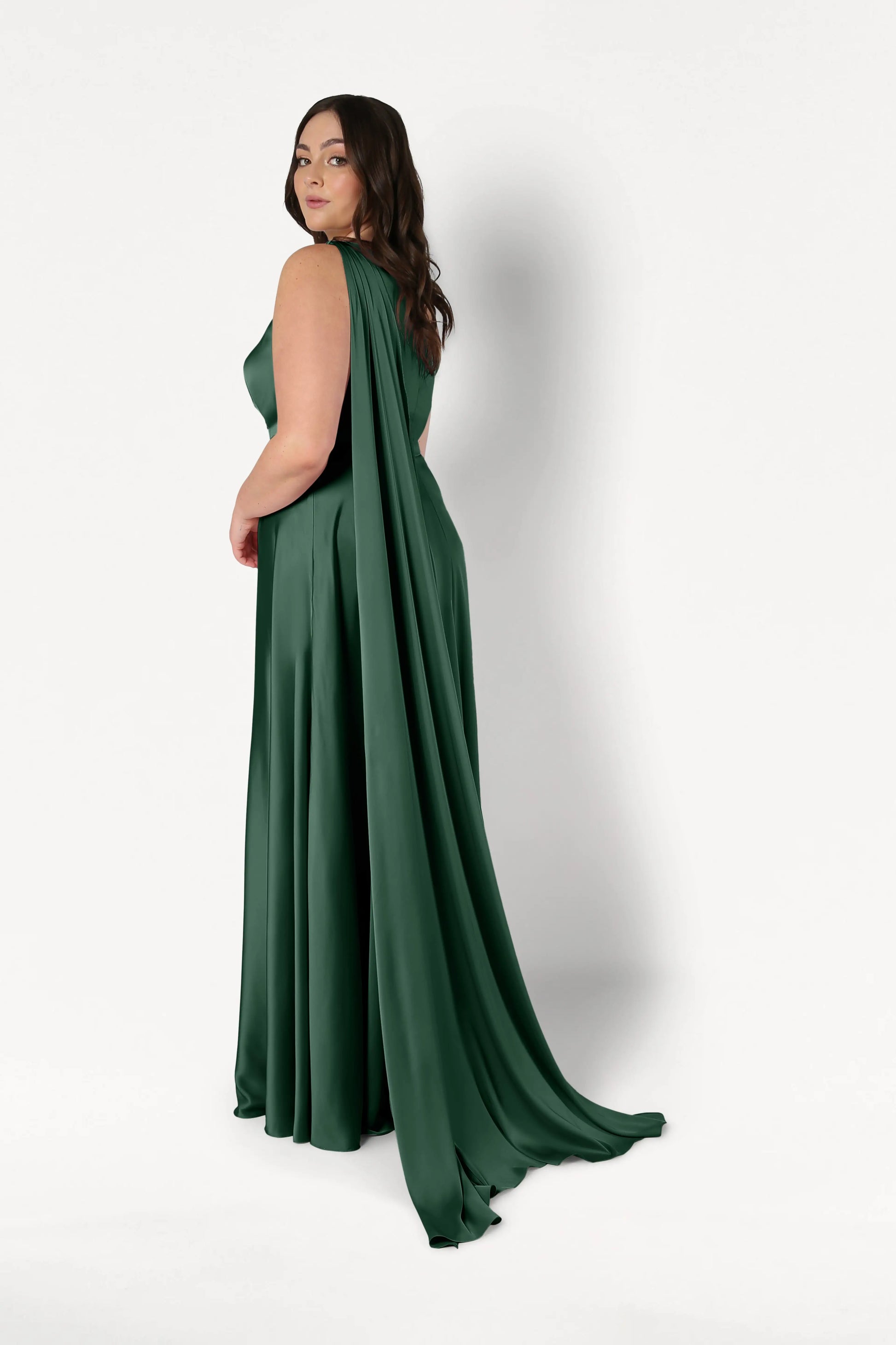 A woman wears the TH&TH Lyra Bridesmaid Dress in Olive Grove Satin, a floor-length, sleeveless gown with flowing shoulder drapes, standing against a plain white background and looking over her shoulder.