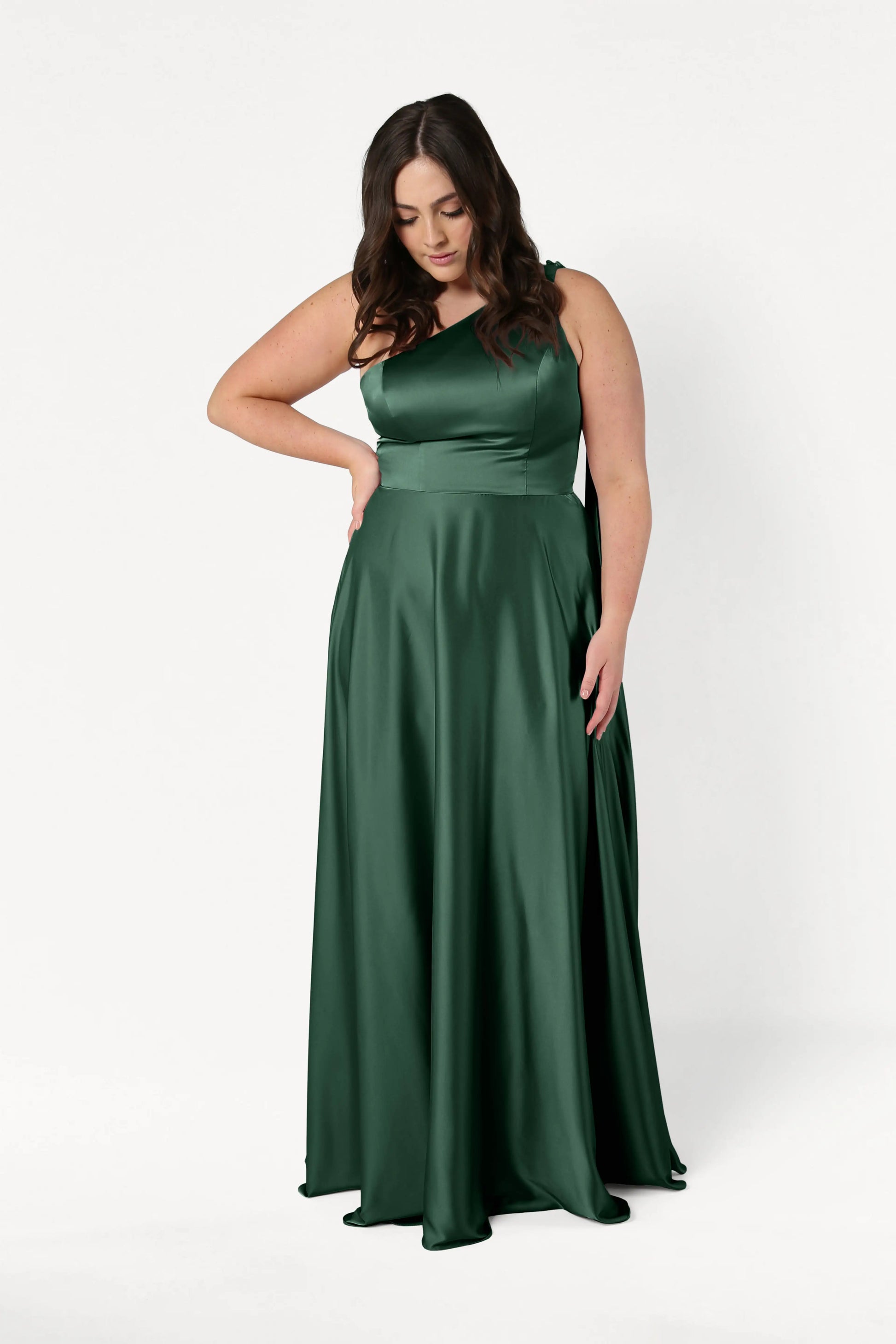 A woman in the Lyra Bridesmaid Dress in Olive Grove Satin by TH&TH stands against a plain white background, looking down with one hand on her hip. This elegant satin gown features a size-inclusive, one-shoulder design.
